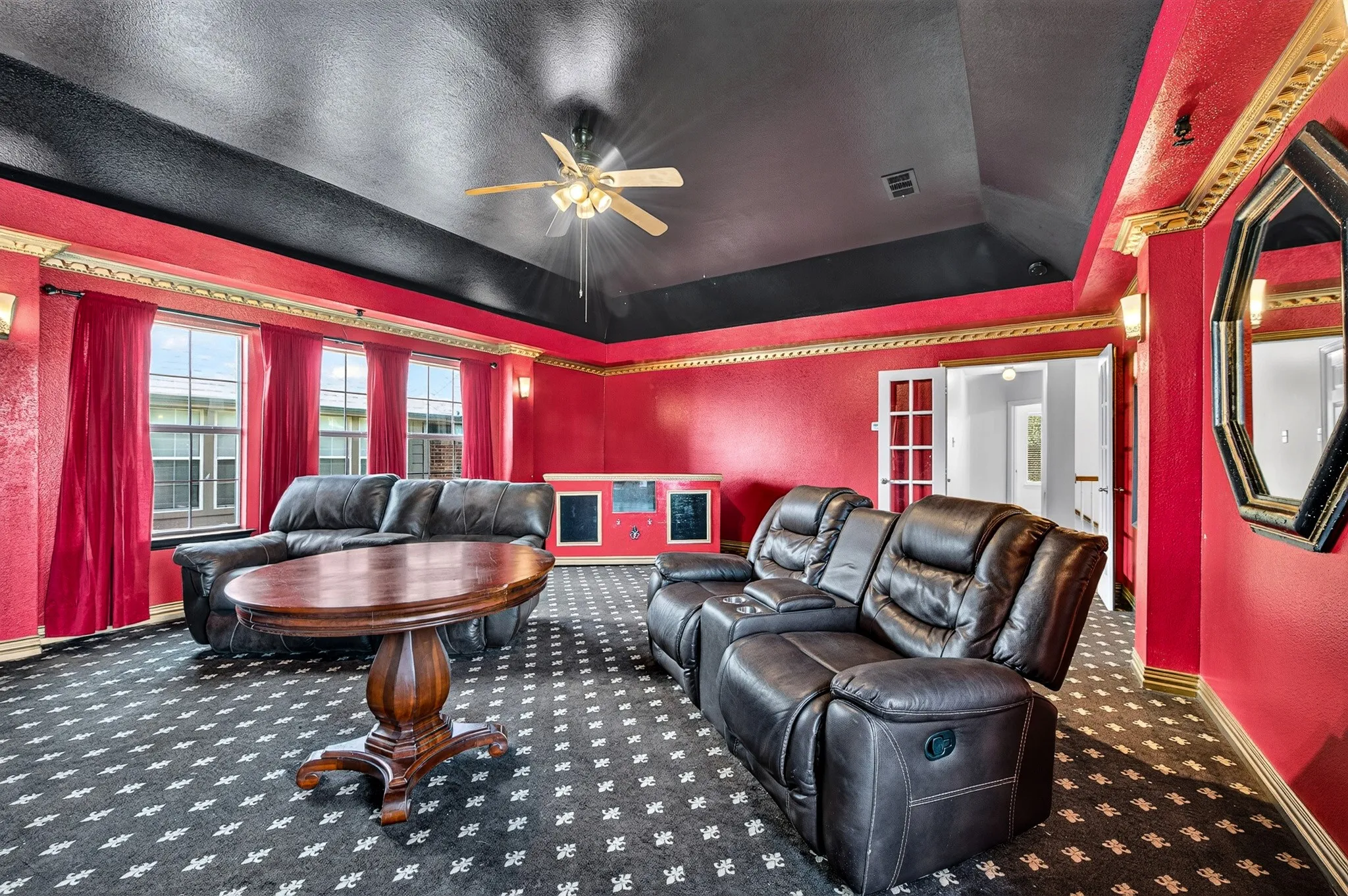 Spacious room featuring patterned carpeting, red textured walls with gold-toned trim, and a black tray ceiling with a ceiling fan