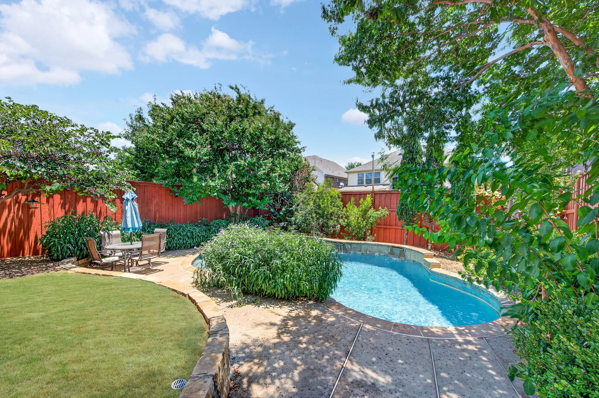 Backyard oasis featuring a clear blue swimming pool, established perimeter landscaping, red-toned privacy fencing, and a manicured lawn area
