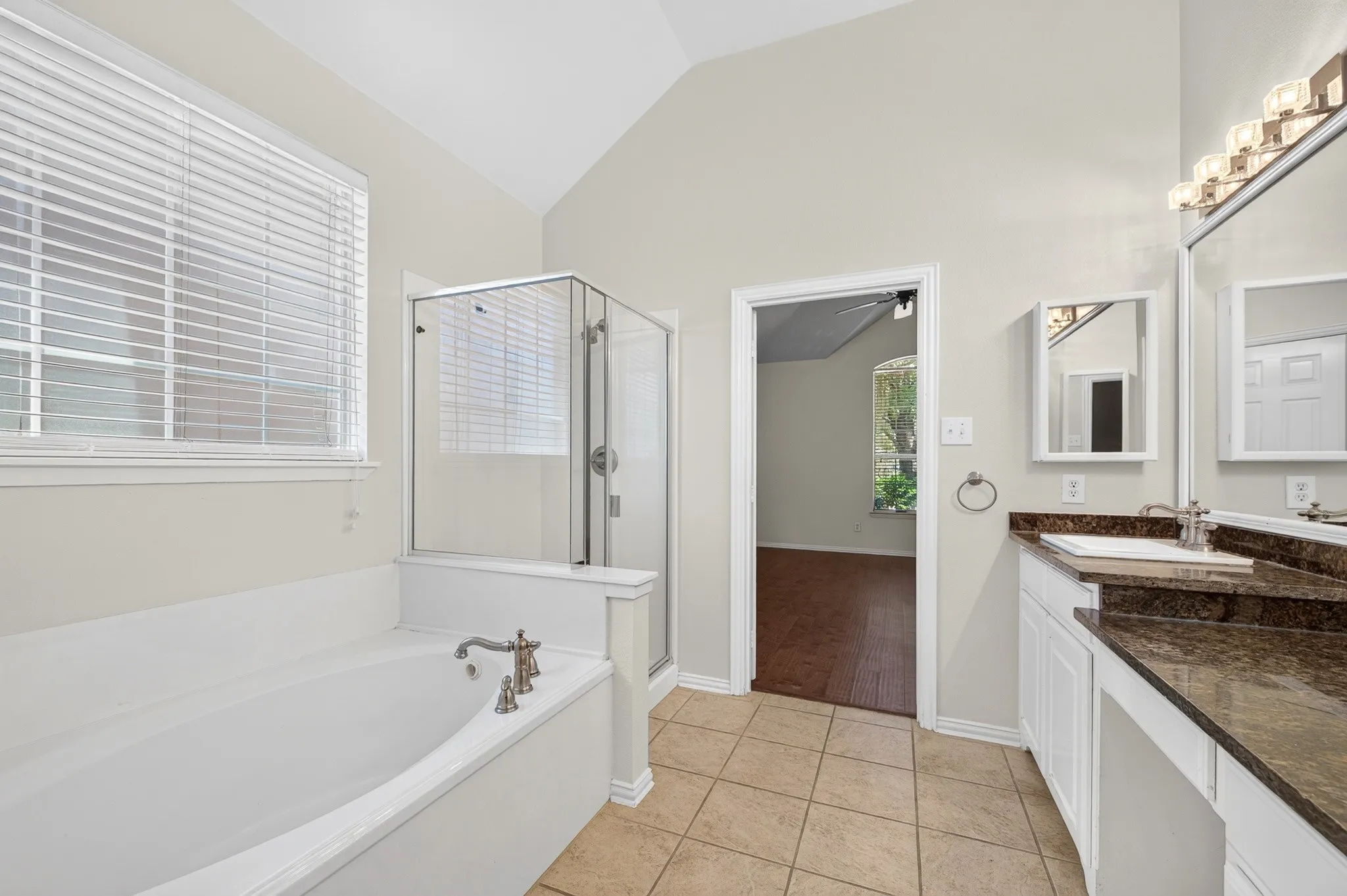Soaking tub with a bronze-finish faucet, an adjacent glass-enclosed shower, a double vanity with a dark speckled countertop, and tiled flooring
