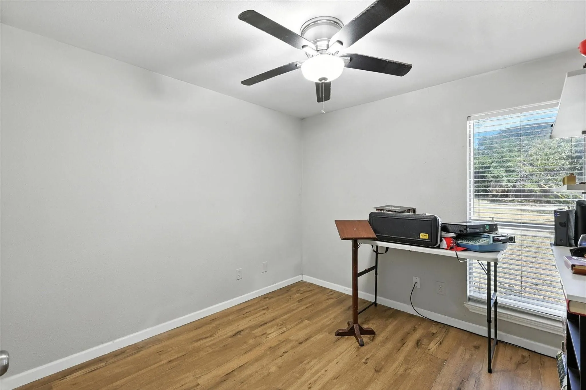 Wood-finish flooring, light gray wall paint, window with blinds, and a ceiling fan with integrated lighting