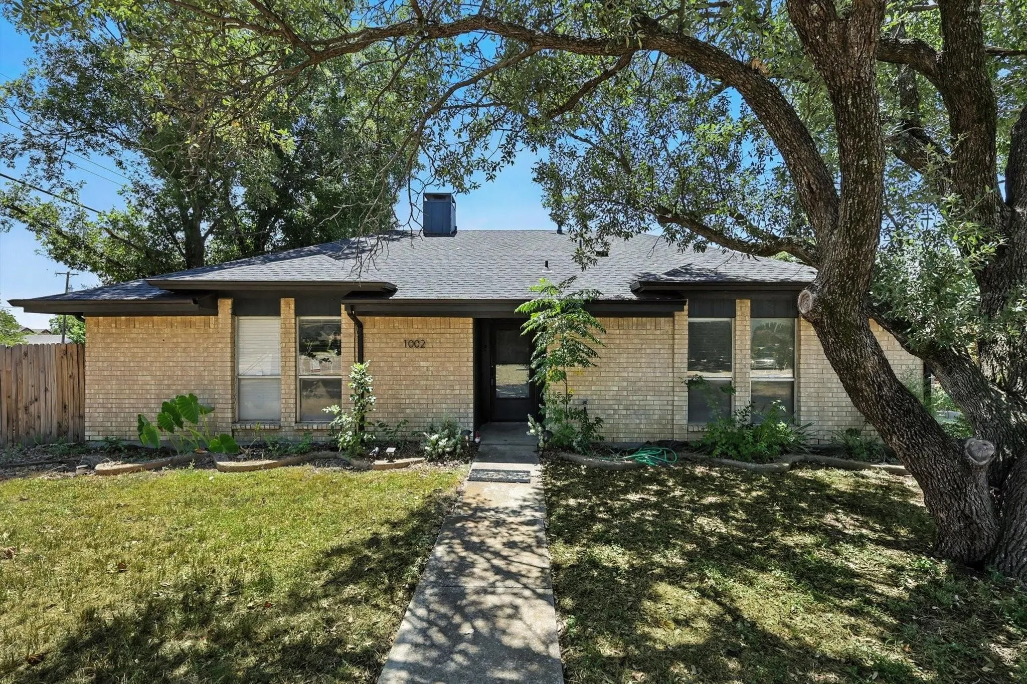Single-story residence featuring light-colored brick siding, a dark shingle roof, a paved walkway, and a mature tree providing natural shade