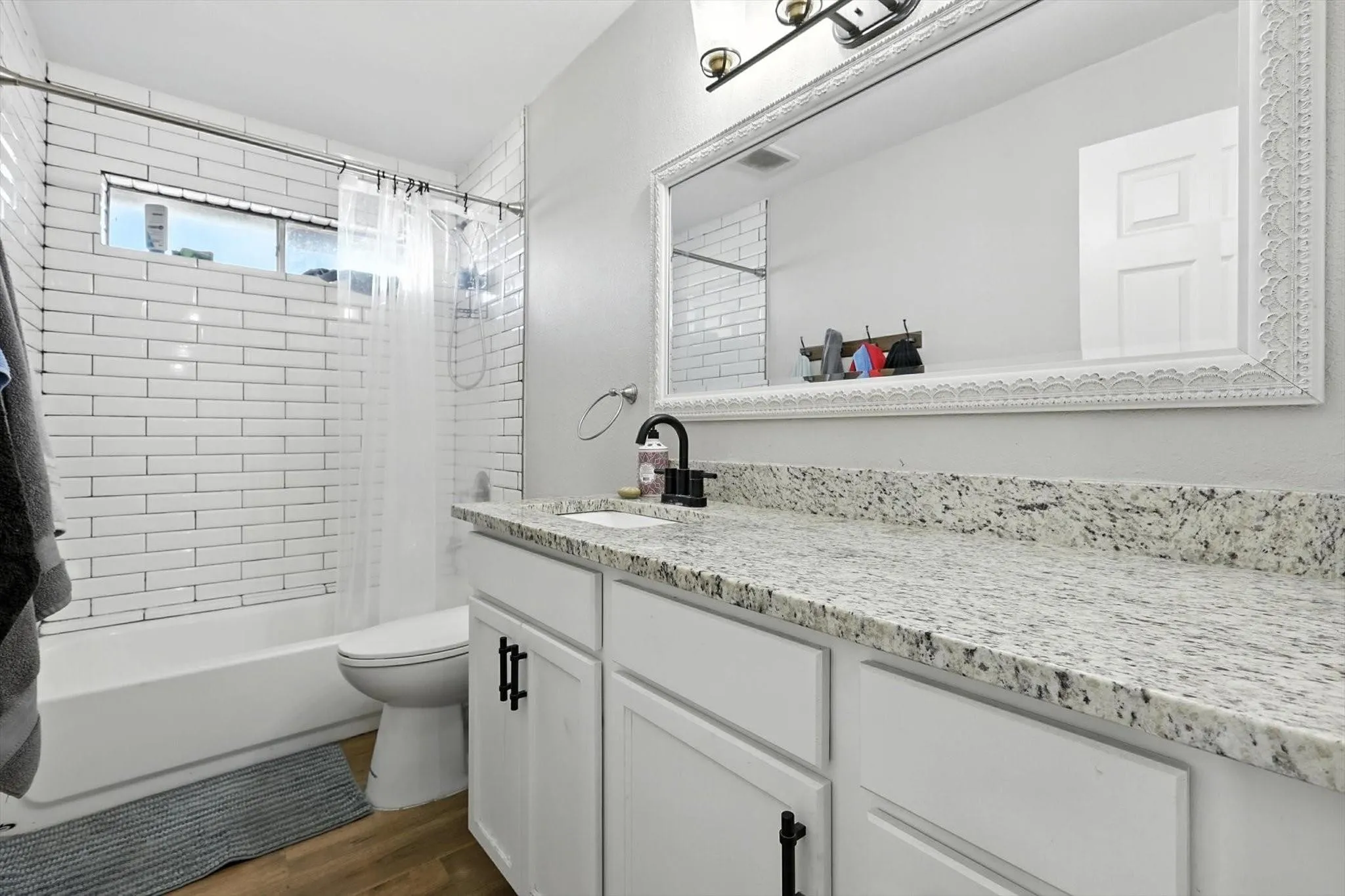 White subway tile shower surround, window above the tub, wood-finish flooring, white vanity with black hardware, and a light-colored speckled countertop