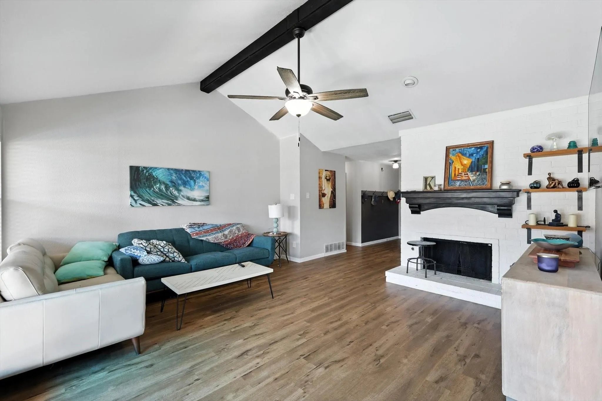 Vaulted ceiling with exposed beam, wood-finish flooring, white brick fireplace with dark wood mantel, ceiling fan, and built-in shelving