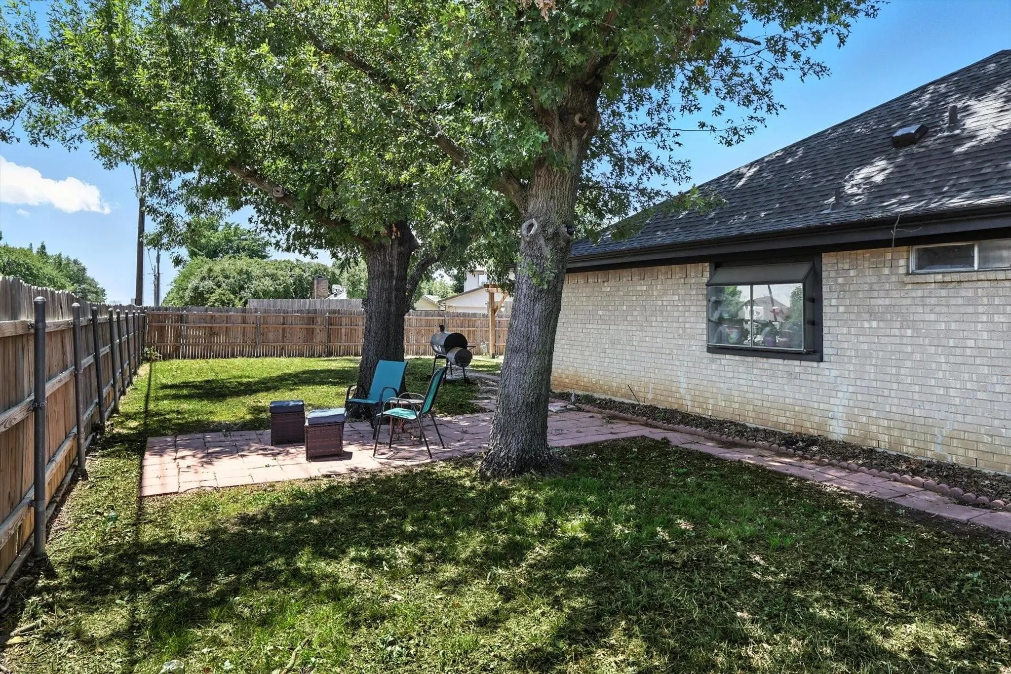 Expansive backyard featuring a manicured lawn, established trees, a brick paver patio, wood fencing, and a white brick exterior