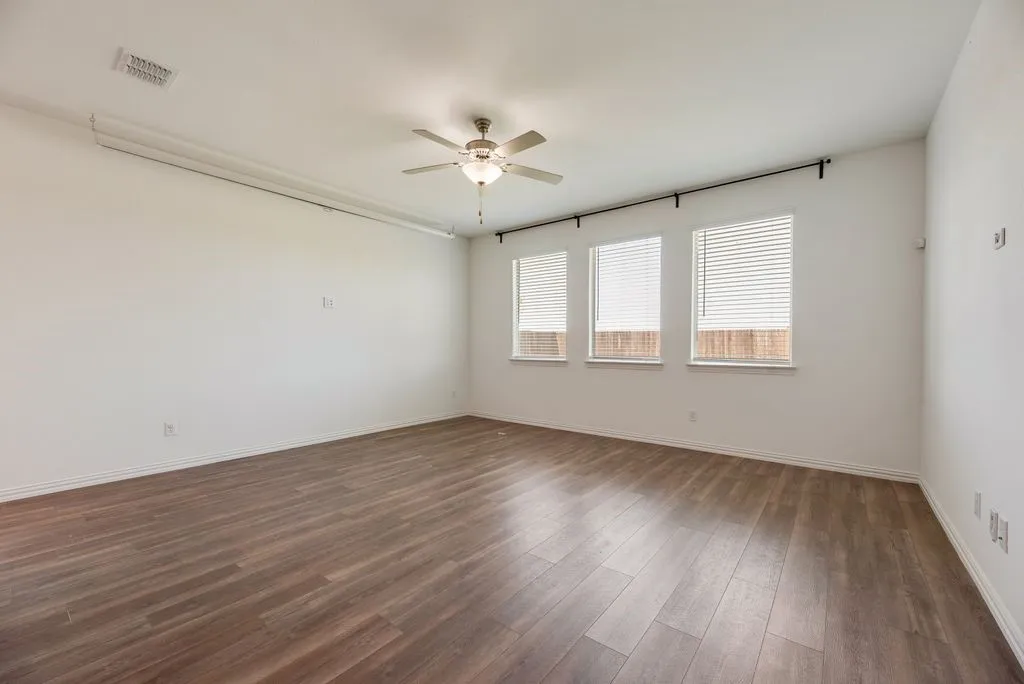 Spacious room featuring wood-finish flooring, three windows with blinds, a ceiling fan with integrated lighting, and a white baseboard trim