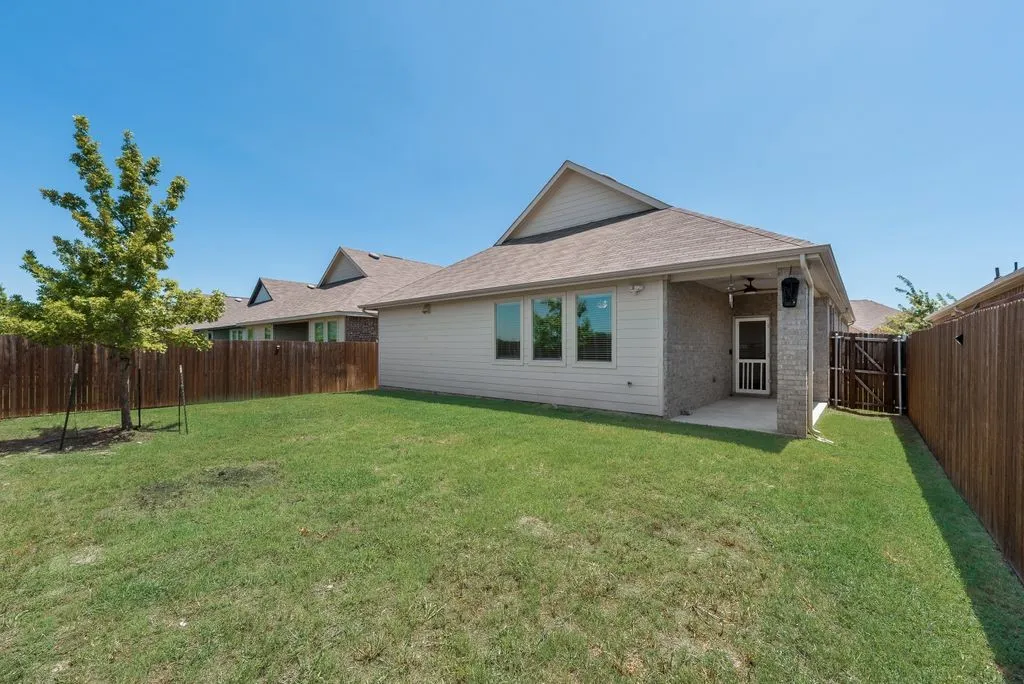 Expansive green lawn with a newly planted tree, a covered patio featuring a ceiling fan and exterior lighting, a full wood fence enclosing the property, and brick and siding exterior finishes