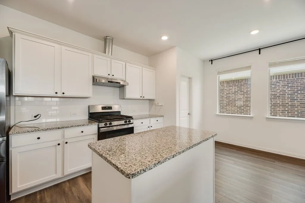 Kitchen featuring an island with granite countertops, white cabinetry with black hardware, stainless steel appliances, a tile backsplash, and wood-finish flooring