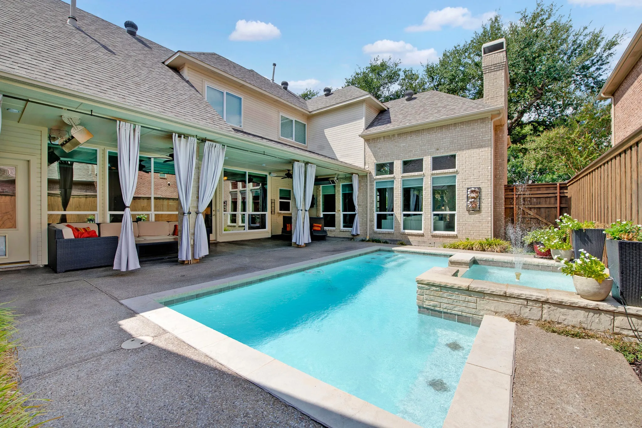 Outdoor swimming pool with integrated spa and water feature, complemented by a covered patio with privacy drapery, and a brick chimney exterior