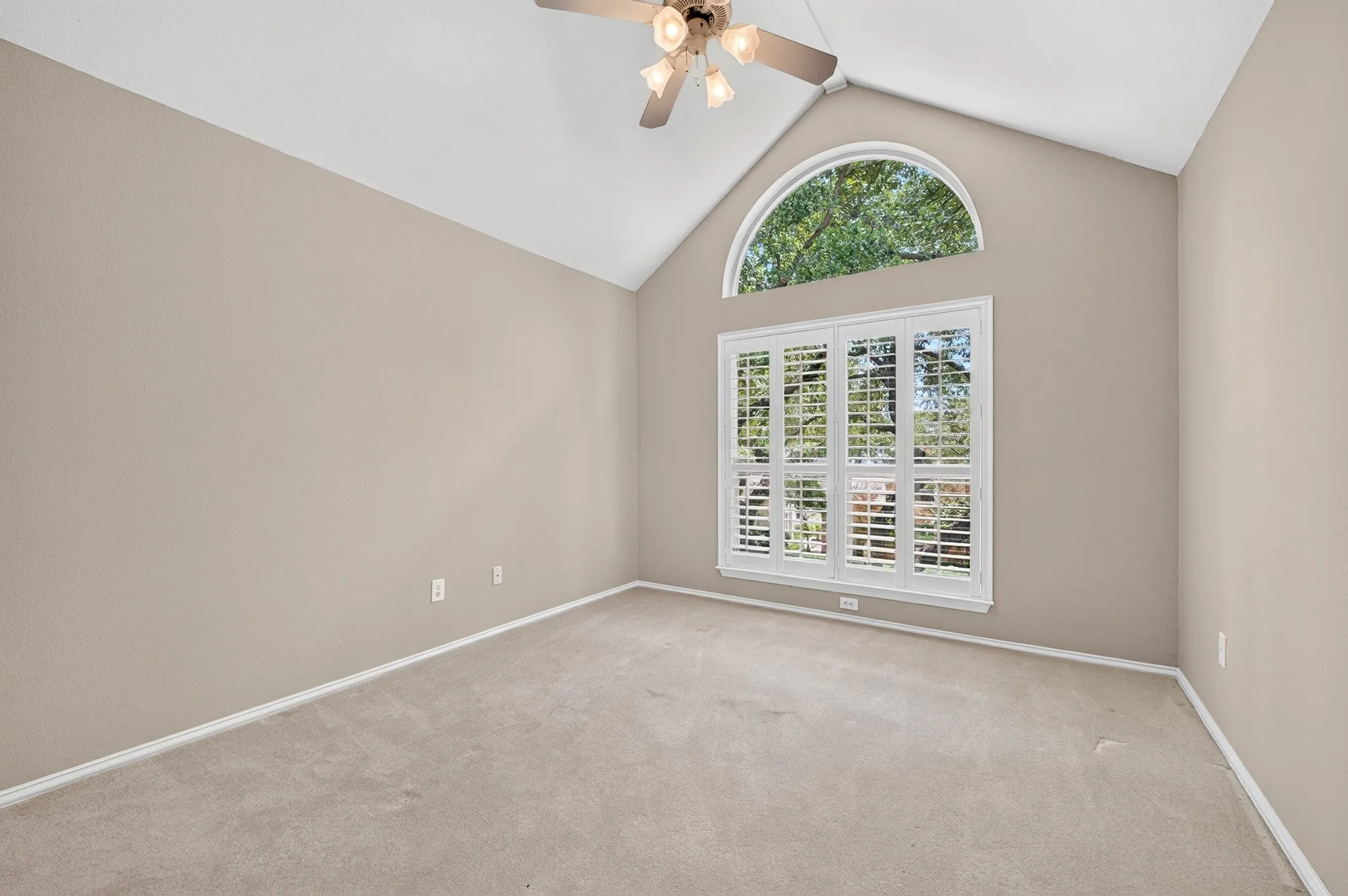 Vaulted ceiling bedroom featuring a large arched window, white plantation shutters, and a ceiling fan with integrated lighting
