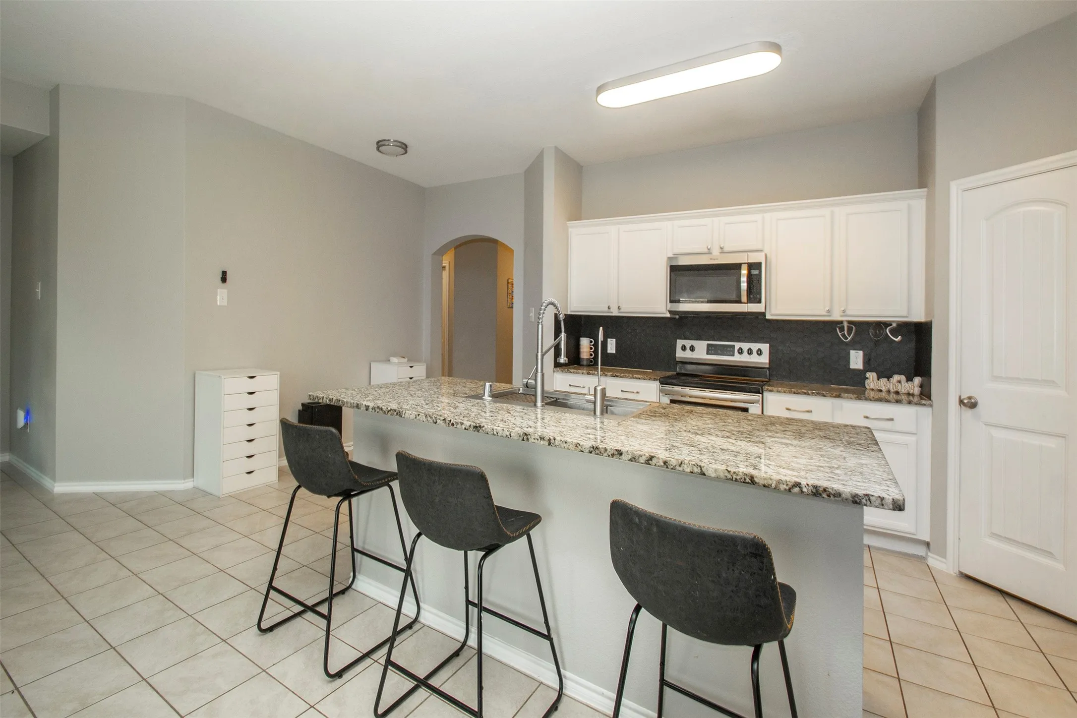 Kitchen featuring a granite-topped island with seating, white cabinetry, stainless steel appliances, dark backsplash, and tile flooring