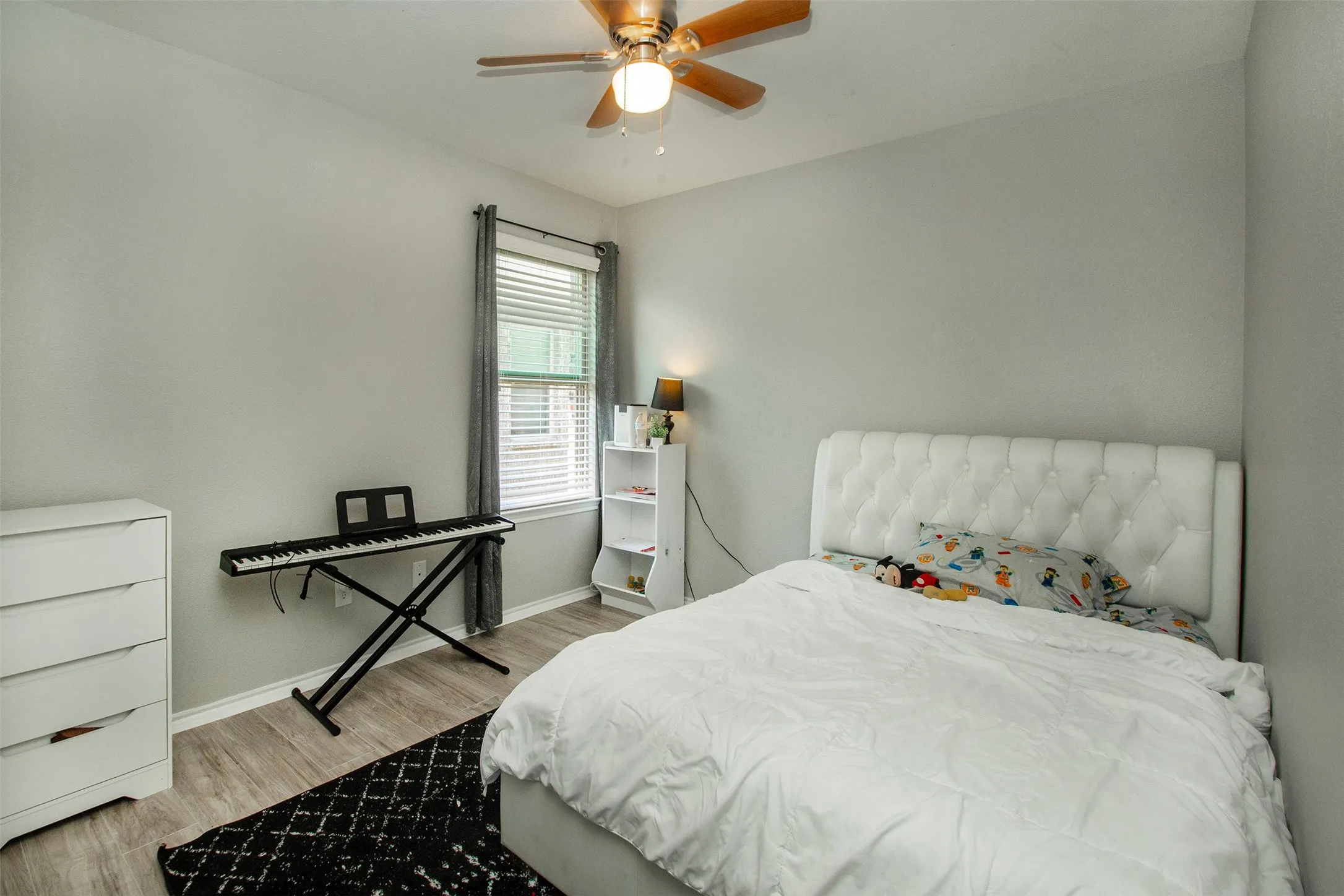 Neutral-toned room featuring wood-finish flooring, a ceiling fan with light fixture, and a window with blinds and drapes