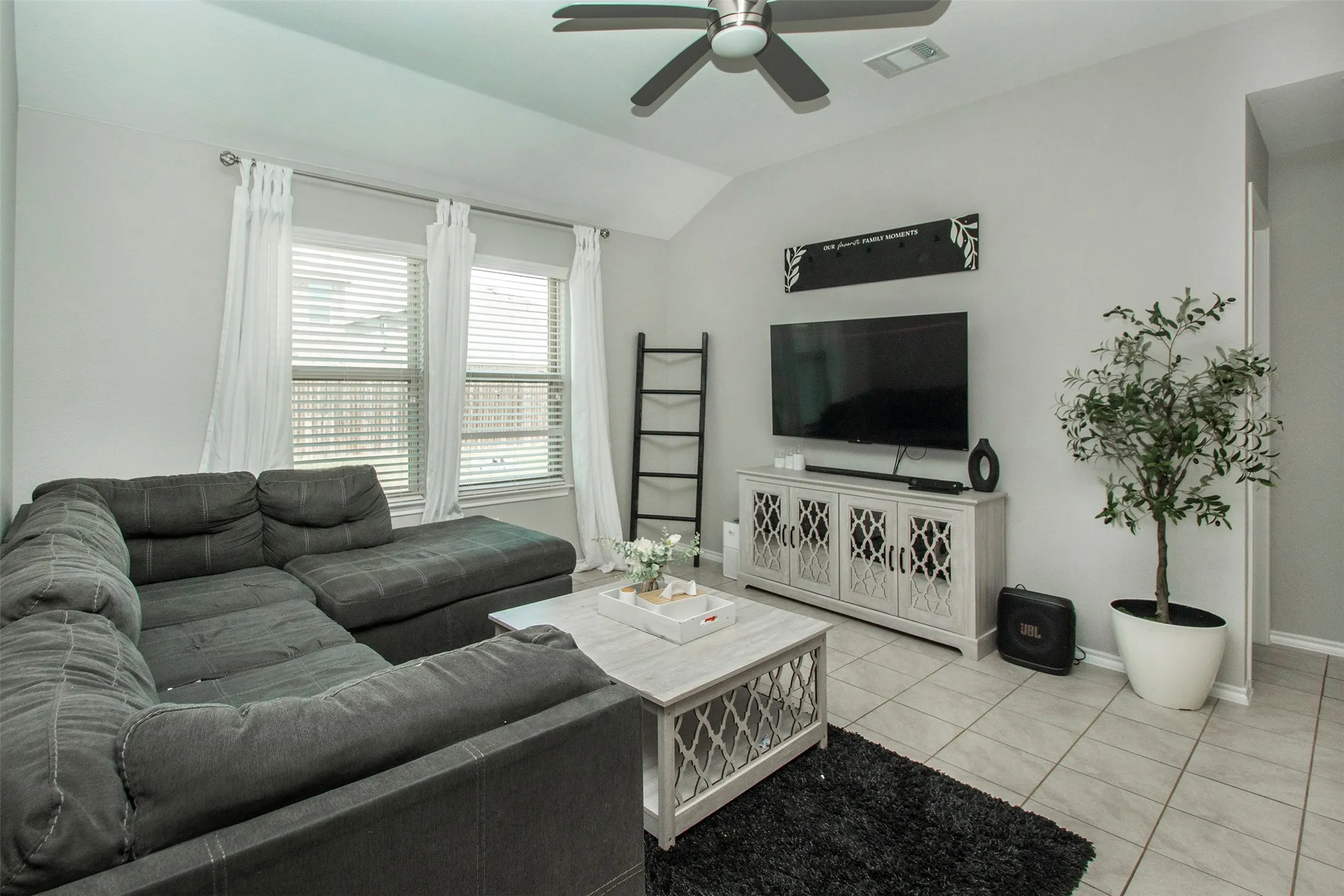 Living area featuring vaulted ceilings, light gray wall paint, multiple windows with blinds, a ceiling fan, and tile flooring