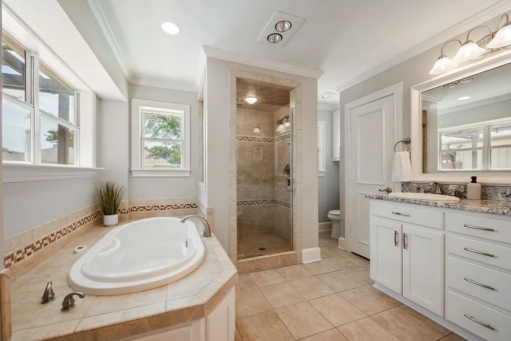 Spacious bathroom featuring a soaking tub, separate tile-surround shower with glass enclosure, dual sink vanity with granite countertop, extensive tile flooring, and multiple windows