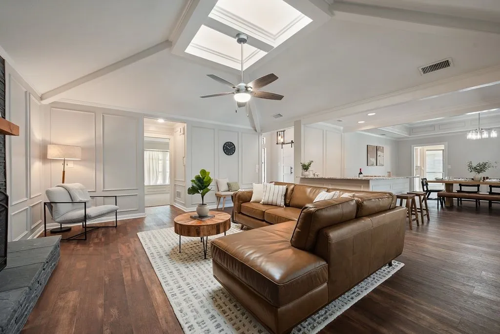 Expansive living area featuring vaulted ceilings with skylights, wood-finish flooring, a stone-clad fireplace with a wood mantel, and wainscoting paneling