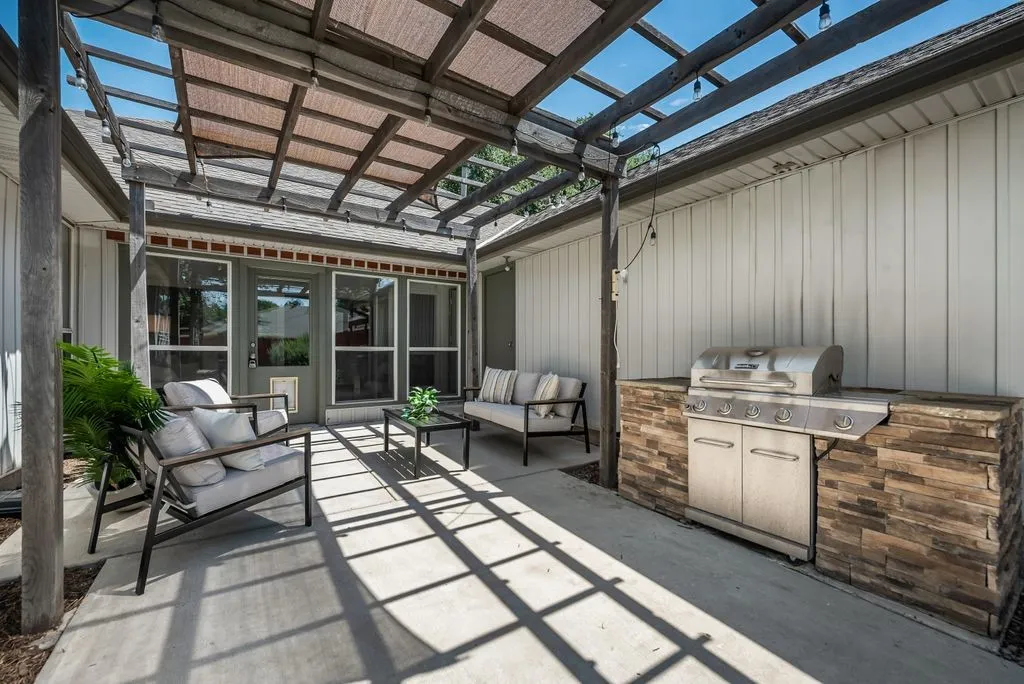 Expansive concrete patio featuring a wood pergola with a sunshade, a built-in outdoor kitchen with stacked stone accents, and exterior glass-paned doors