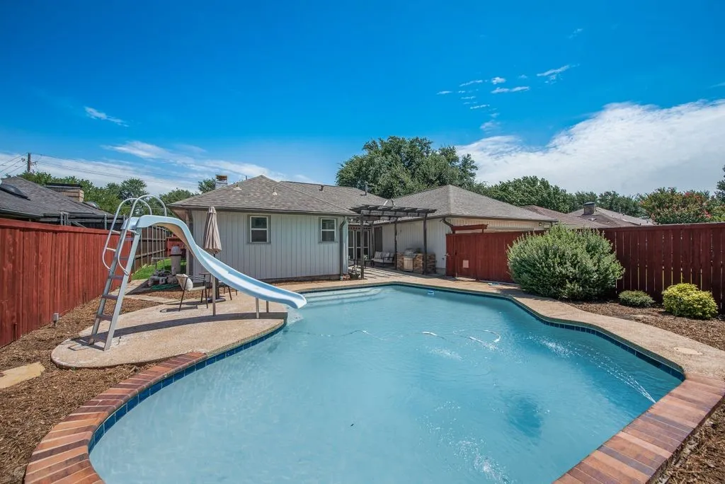 Fenced backyard featuring a swimming pool with a slide, concrete decking, and a covered patio area