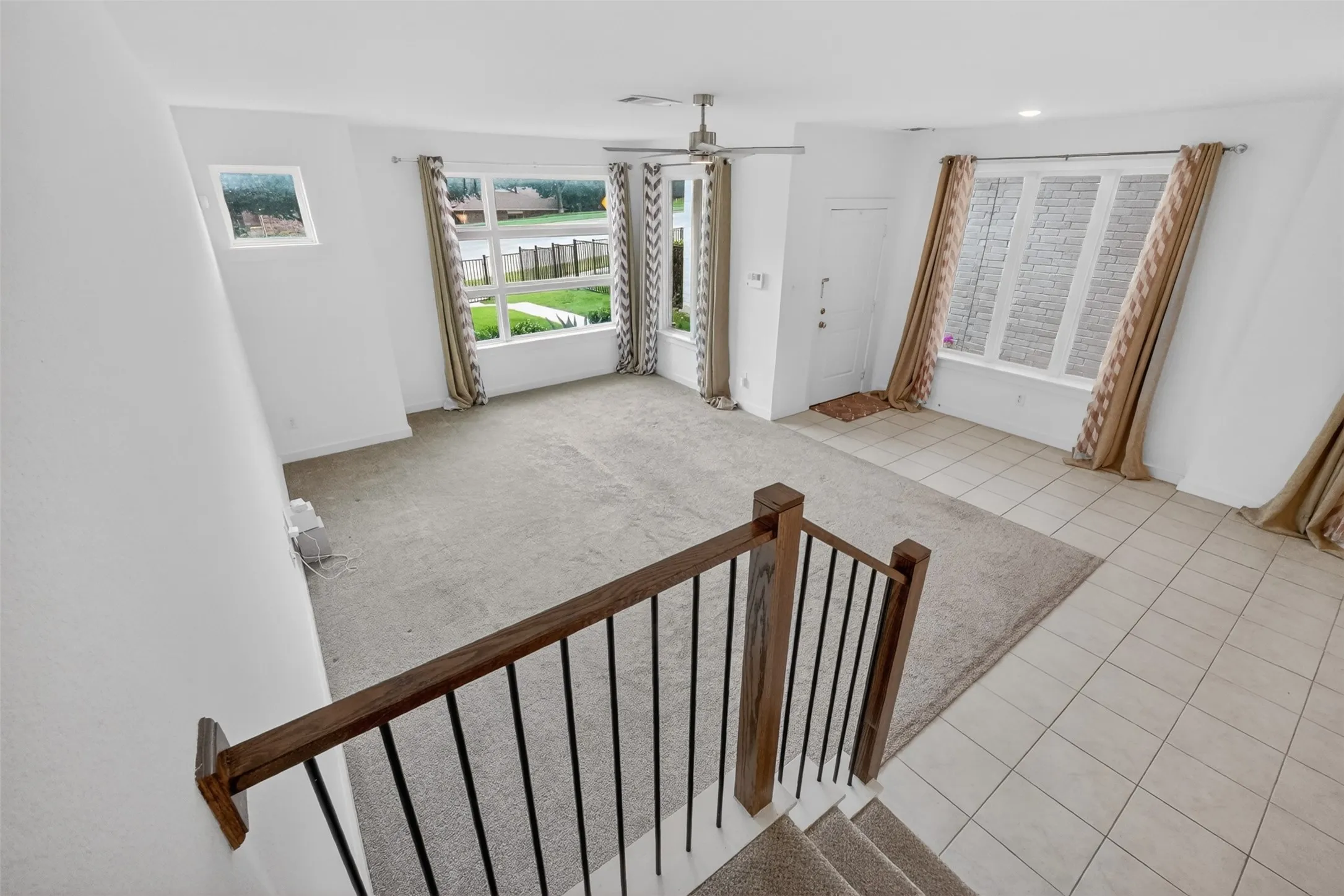Spacious living area featuring a ceiling fan, multiple windows with neutral drapes, and a combination of carpet and tile flooring