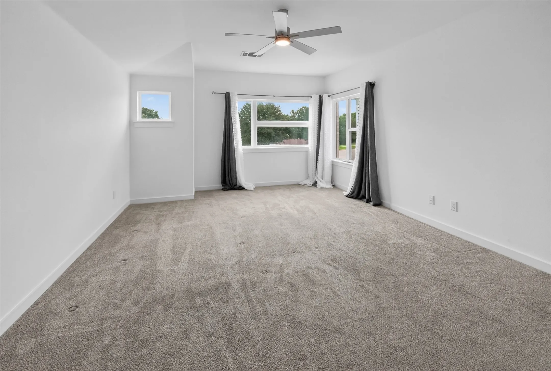 Spacious room featuring neutral carpeting, white walls, multiple windows, a ceiling fan with light, and white baseboards
