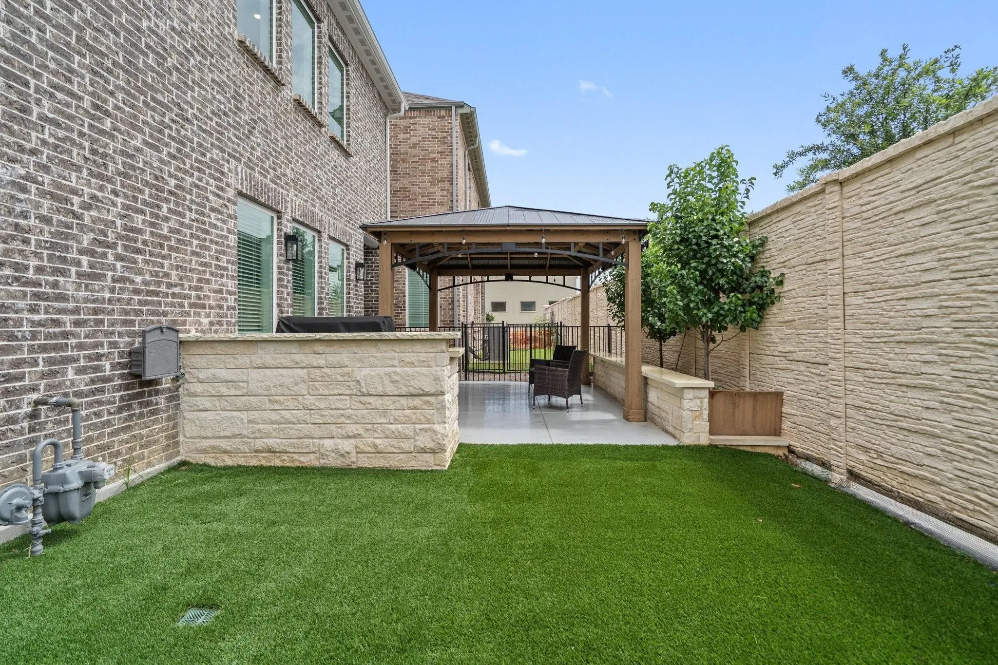 Backyard featuring artificial turf, a stone-faced outdoor kitchen base, and a covered patio with a metal gazebo structure