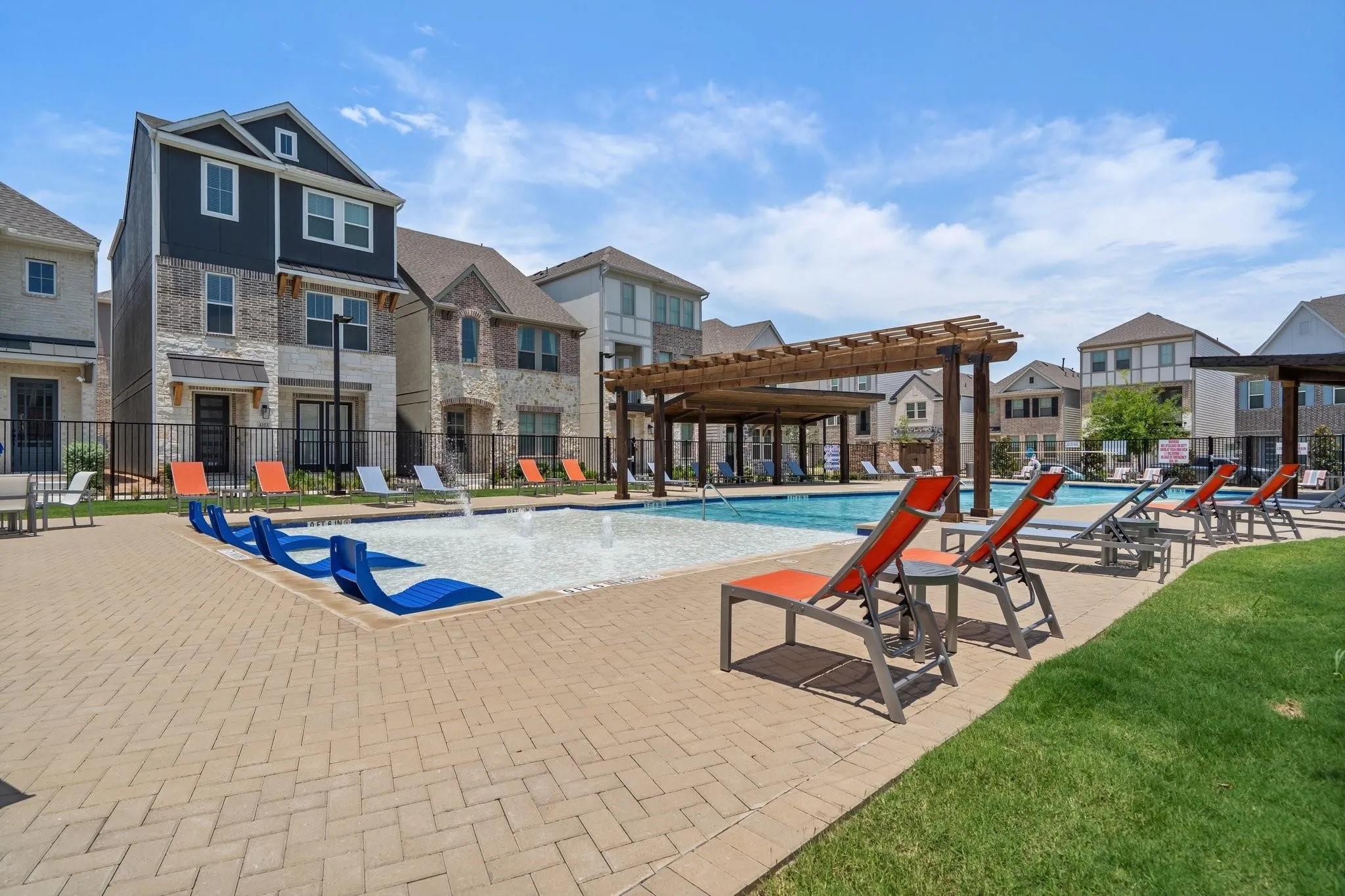 Resort-style community pool area featuring a paver deck, multiple lounge chairs, in-pool seating, and shaded pergolas, bordered by manicured turf and architectural townhomes