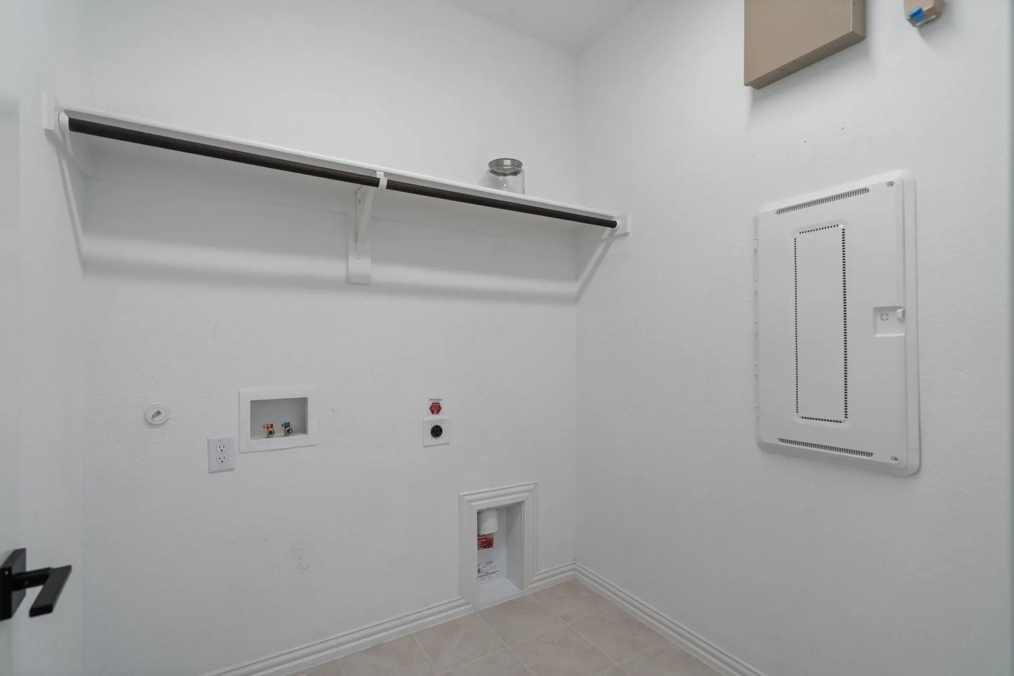 Dedicated laundry area featuring a built-in shelf and hanging rod, electrical outlet, and utility hookups, all complemented by light-toned tile flooring and white baseboards