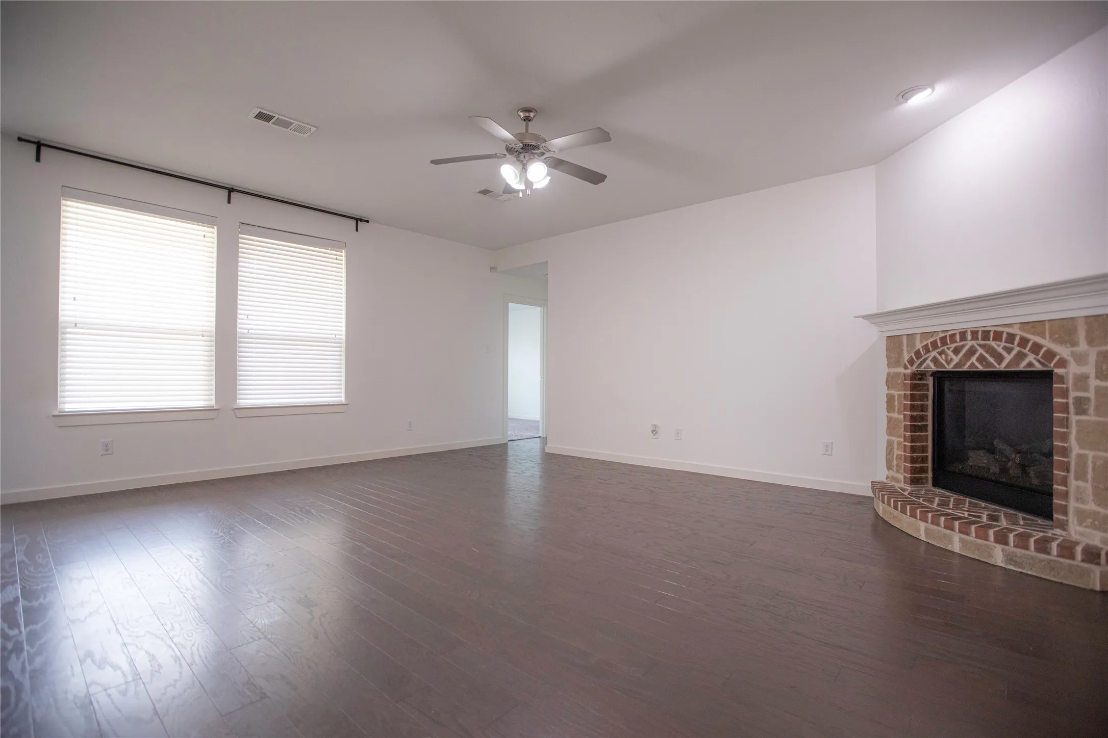 Spacious living area featuring wood-finish flooring, a corner fireplace with brick and stone surround, dual windows, and an overhead ceiling fan