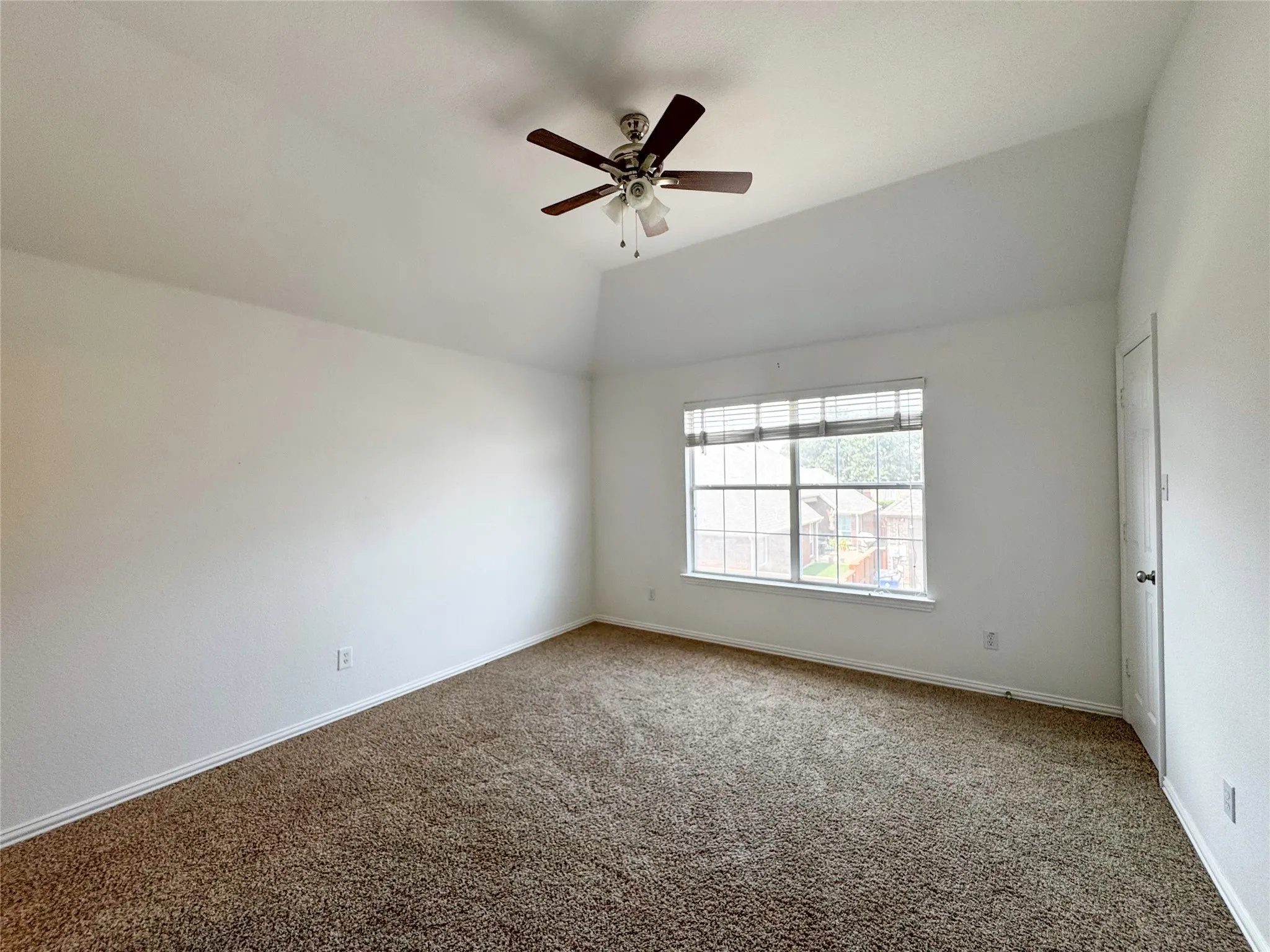 Vaulted ceiling, ceiling fan with light fixture, large window with blinds, brown carpeting, and white baseboards