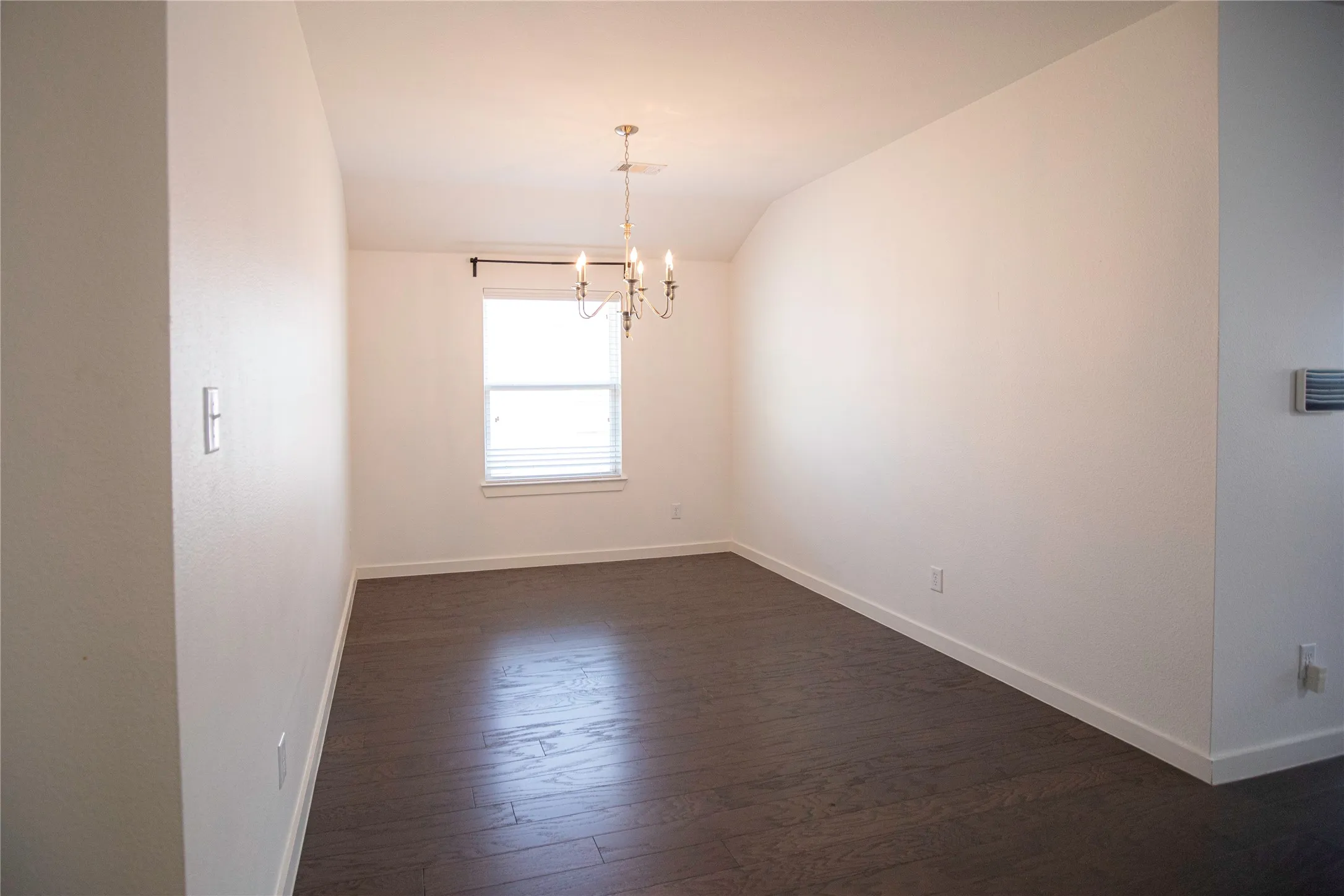 Bright and spacious room featuring pristine white walls, dark wood-finish flooring, a large window with natural light, and a modern pendant light fixture suspended from the ceiling