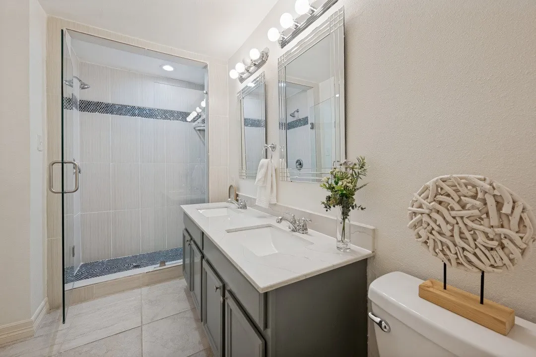Bathroom featuring a dual-sink vanity with a light countertop, two framed mirrors, and a glass-enclosed shower with tile surround and dark accent mosaic