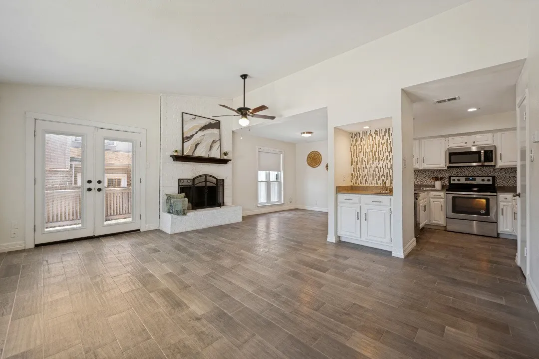 Open-concept living area featuring wood-finish tile flooring, a white brick fireplace with a wood mantle, and double French doors opening to an outdoor deck