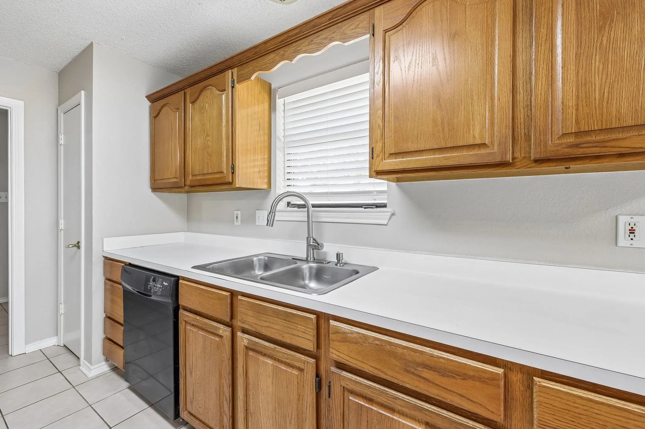 Kitchen featuring wood cabinetry with decorative valance, white countertops, double basin stainless steel sink with pull-down faucet, and a black built-in dishwasher