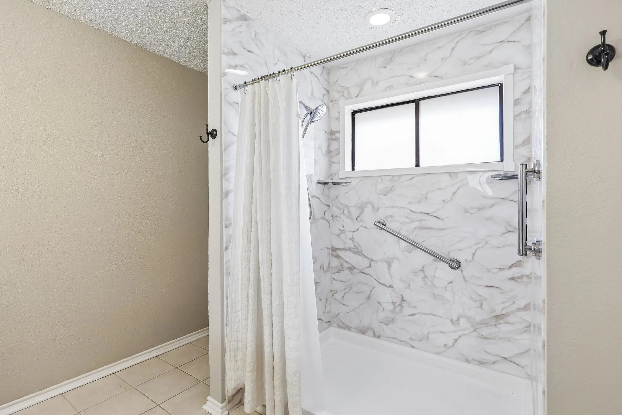 Bathtub with a shower enclosure featuring stone-look wall panels, a frosted window, and chrome grab bars