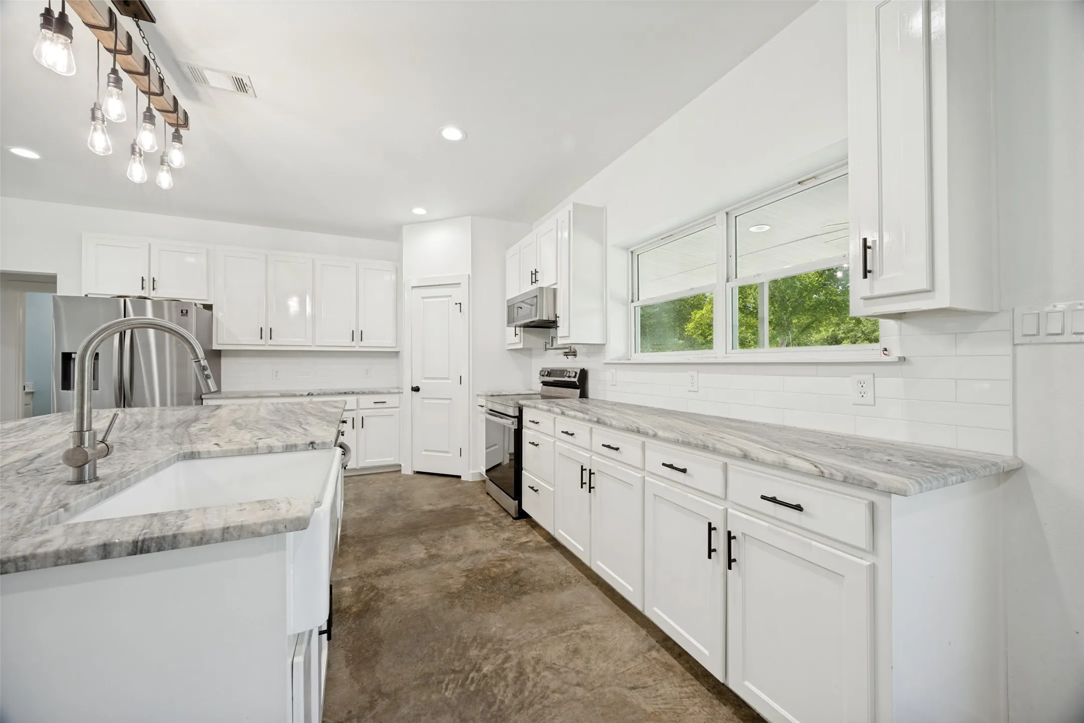 Bright kitchen featuring white shaker cabinetry, stainless steel appliances, a farmhouse sink with a gooseneck faucet, light-toned stone countertops, and a white subway tile backsplash