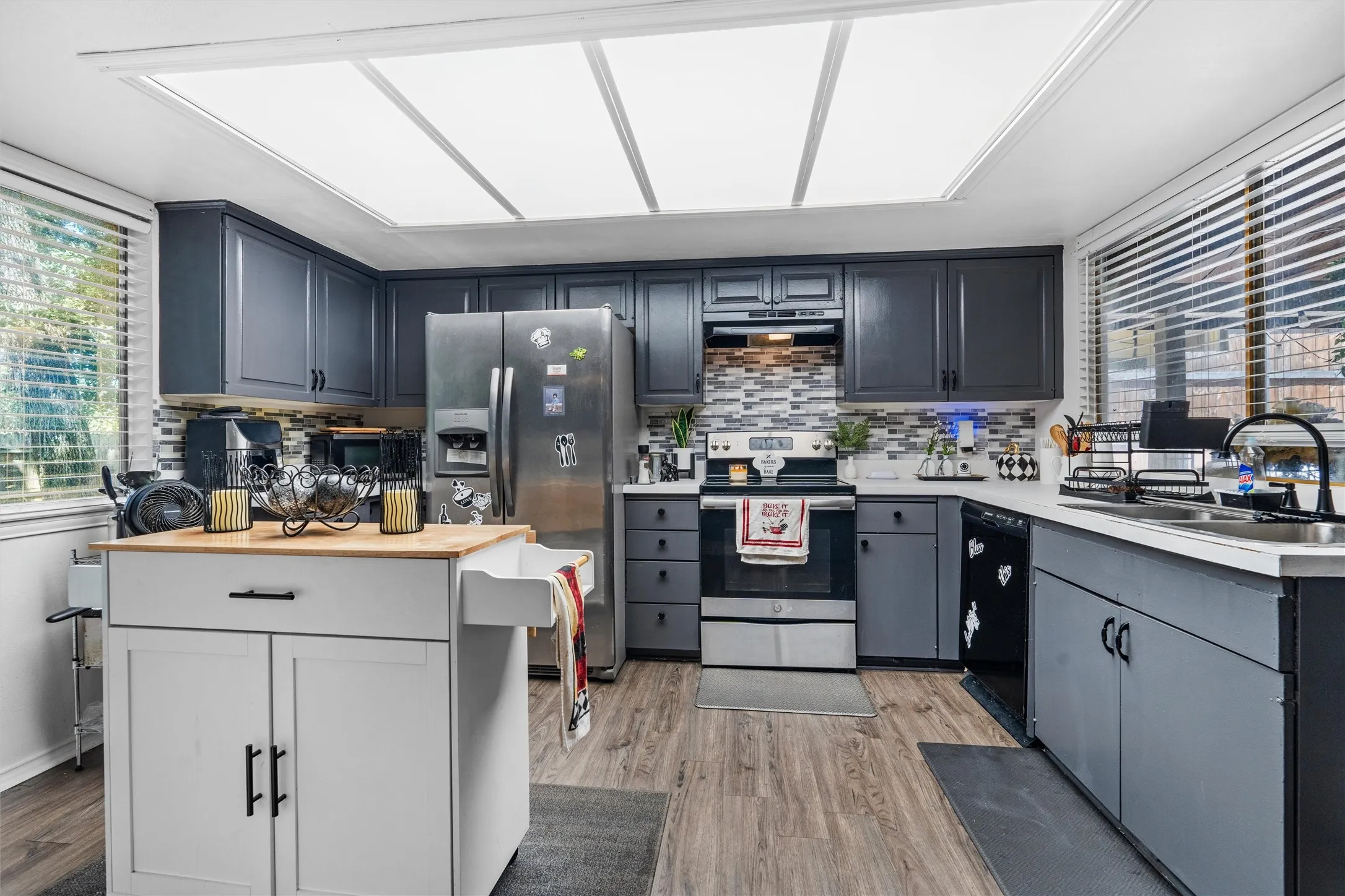 Kitchen featuring gray cabinetry, stainless steel appliances, a tile backsplash, wood-finish flooring, and a large ceiling light fixture