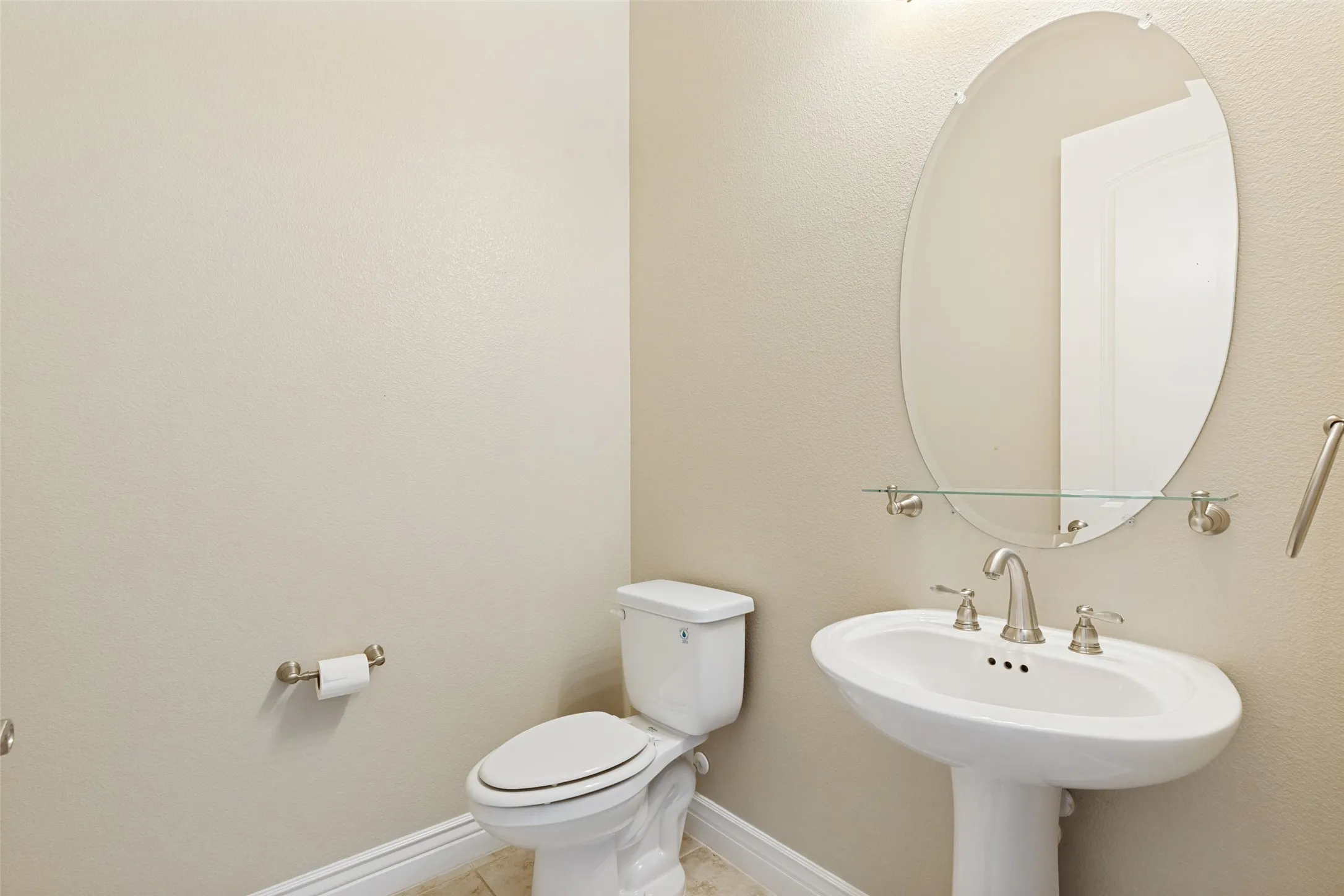 Pedestal sink with brushed nickel hardware, oval frameless mirror with a glass shelf, and a white toilet