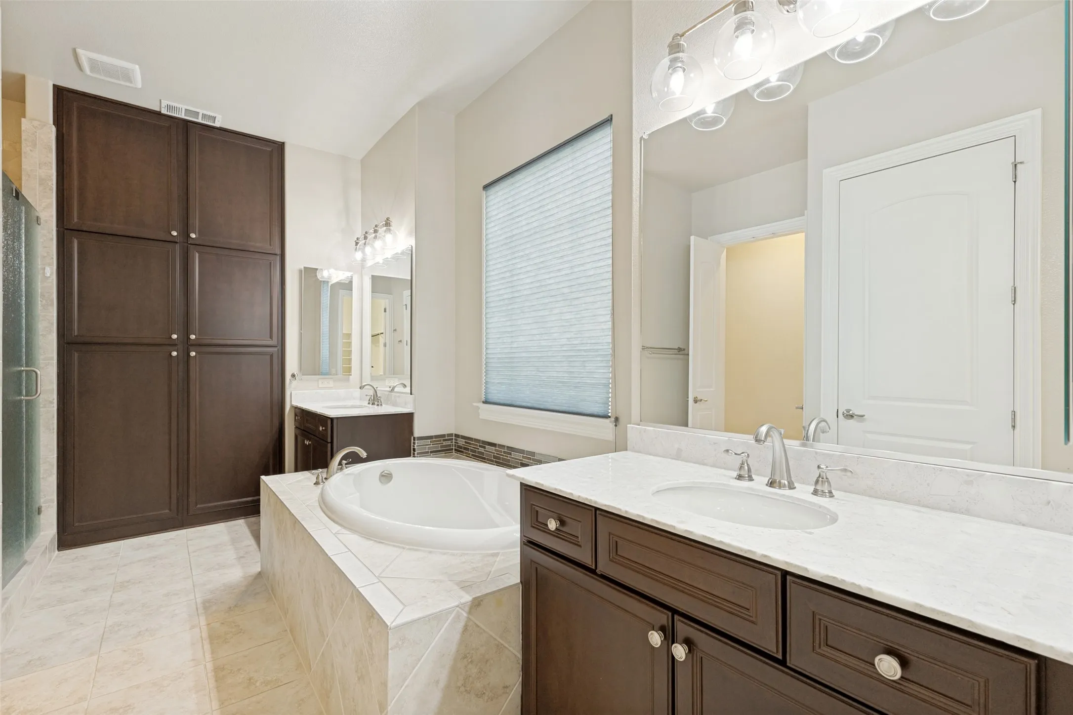 Spacious bathroom featuring a built-in soaking tub with tiled surround, dual vanities with white countertops, dark wood-finish cabinetry, and a large built-in storage unit
