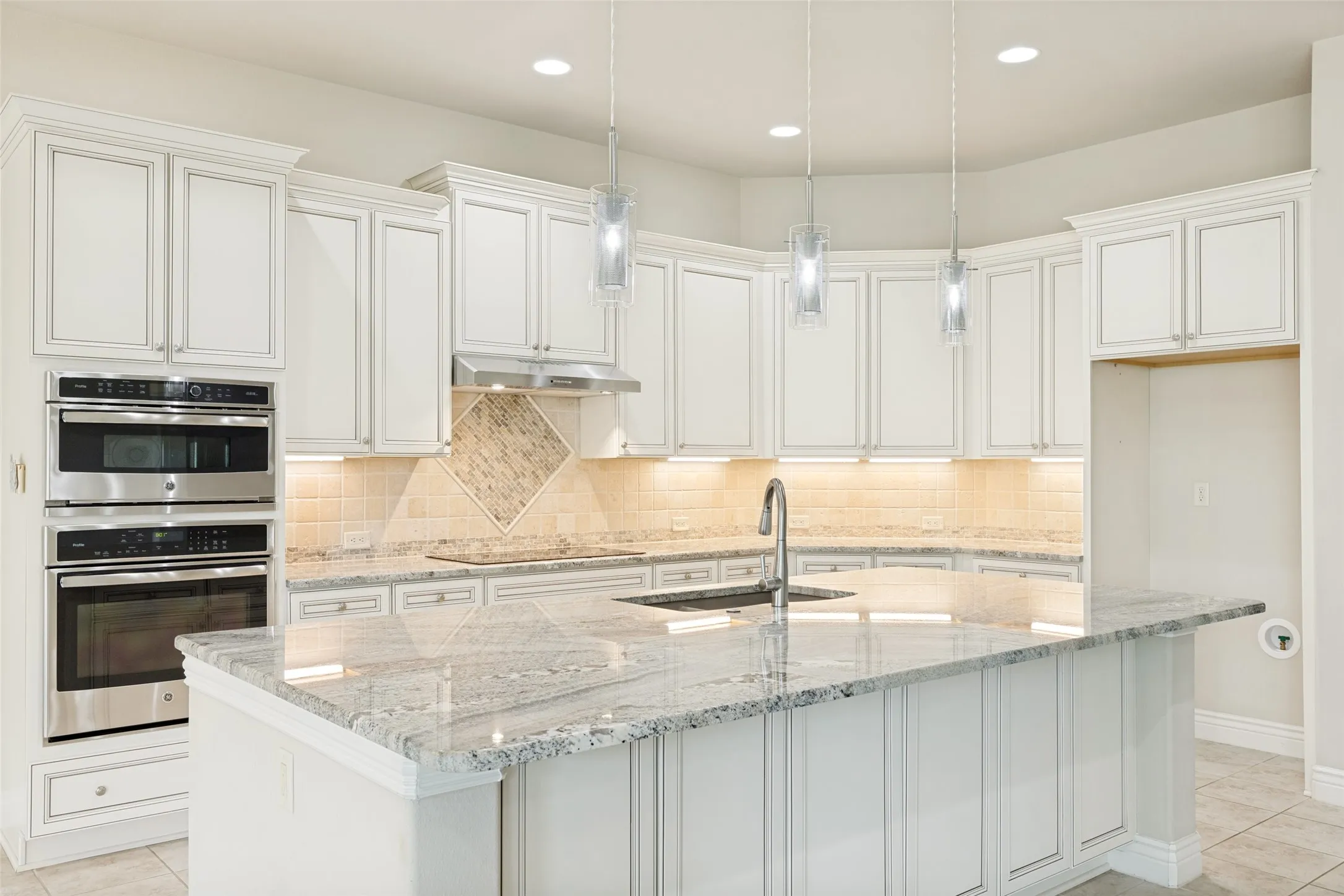 Kitchen featuring a large island with a built-in sink, white cabinetry with crown molding, stainless steel double ovens, recessed lighting, and pendant lights
