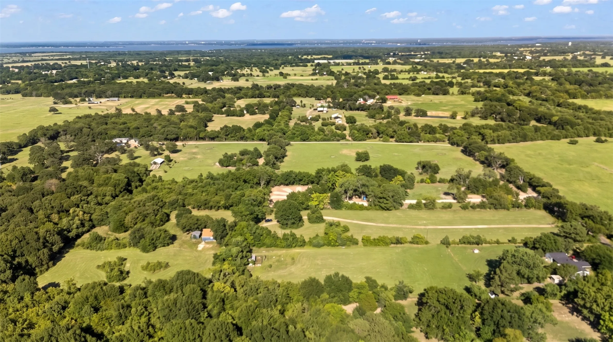 Expansive rural landscape featuring extensive green pastures, abundant mature trees, multiple agricultural structures, and a distant body of water