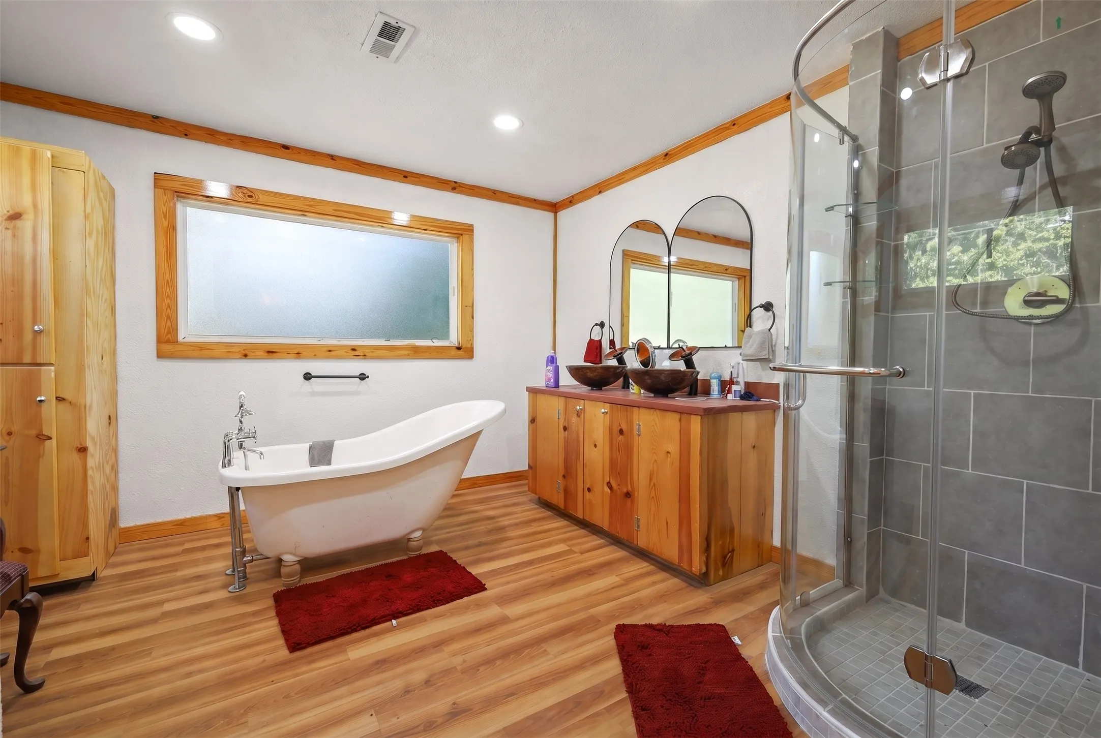 Spacious bathroom featuring a freestanding clawfoot tub, a dual vanity with vessel sinks, a glass-enclosed shower, wood-finish flooring, and natural wood trim accents