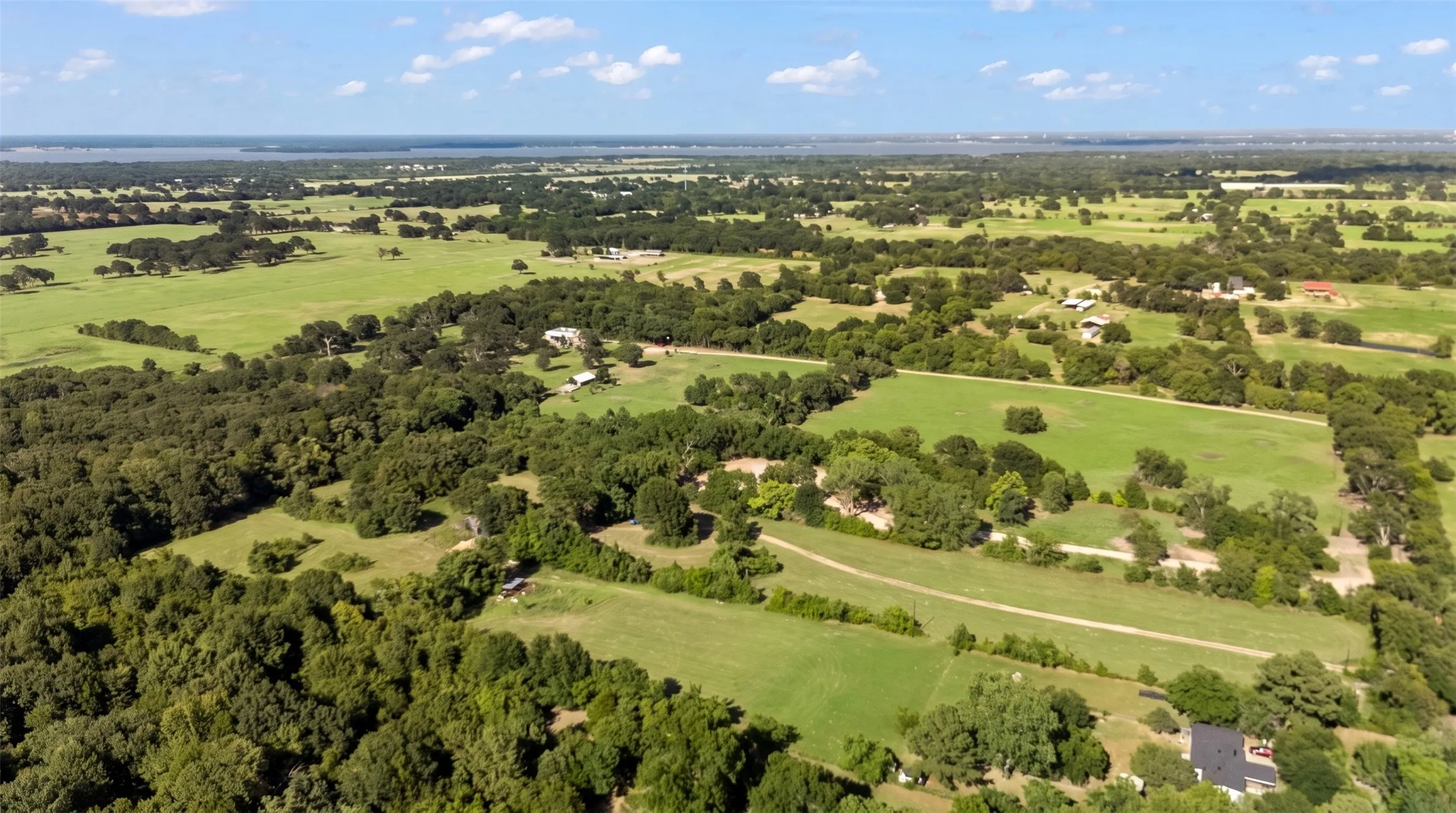 Expansive rural landscape featuring extensive green pastures, mature tree coverage, and a distant body of water