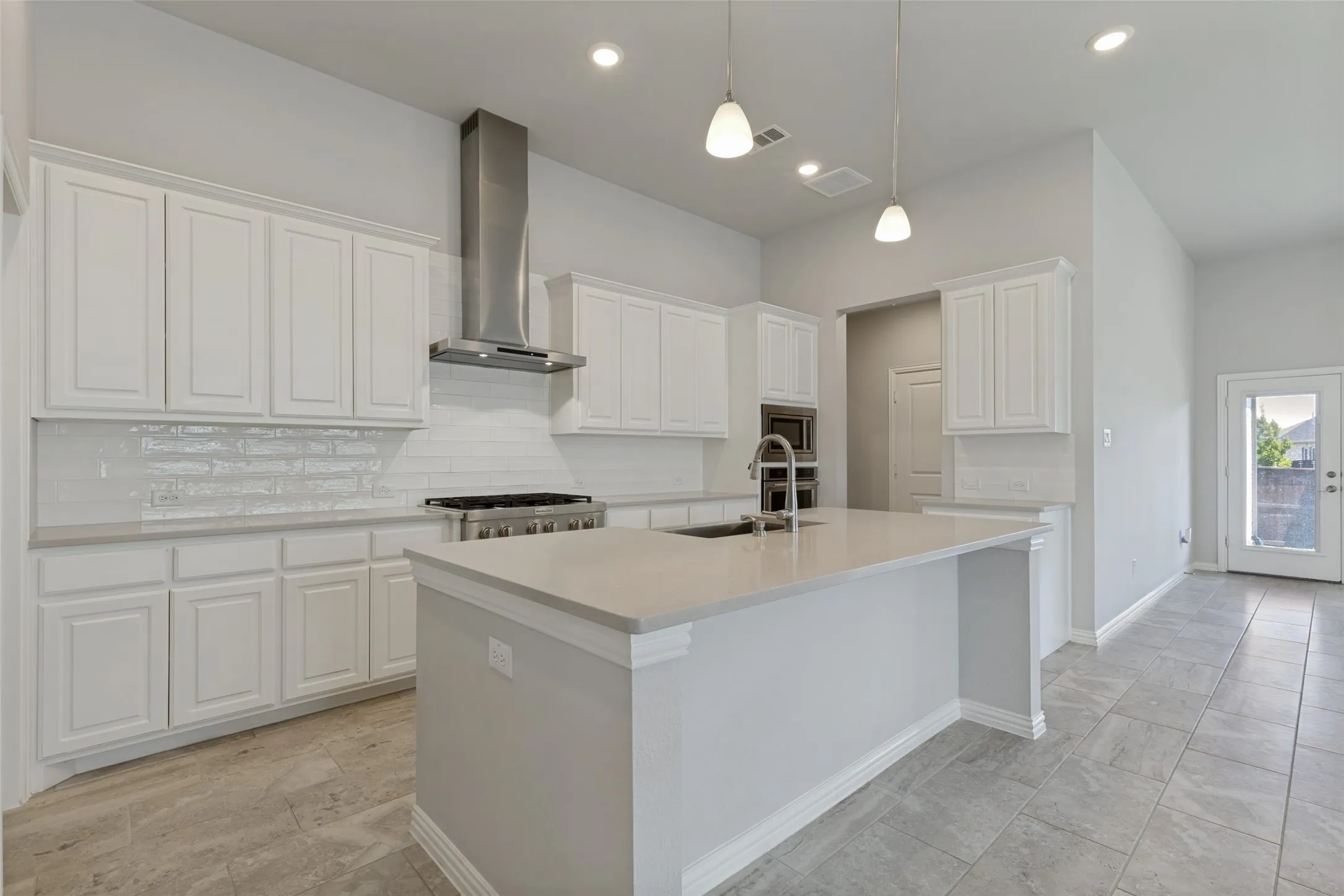 Bright kitchen featuring a central island with an undermount sink, white shaker cabinetry, a subway tile backsplash, a stainless steel range hood, and tile flooring