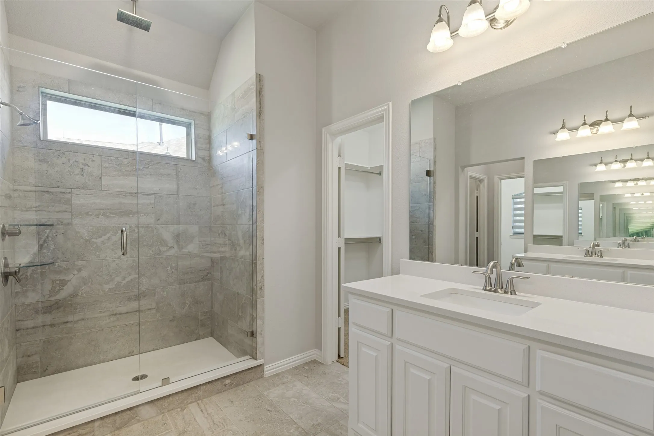 Spacious bathroom featuring a glass-enclosed shower with stone-look tile, transom window, dual vanity with white cabinetry, and a large wall-mounted mirror