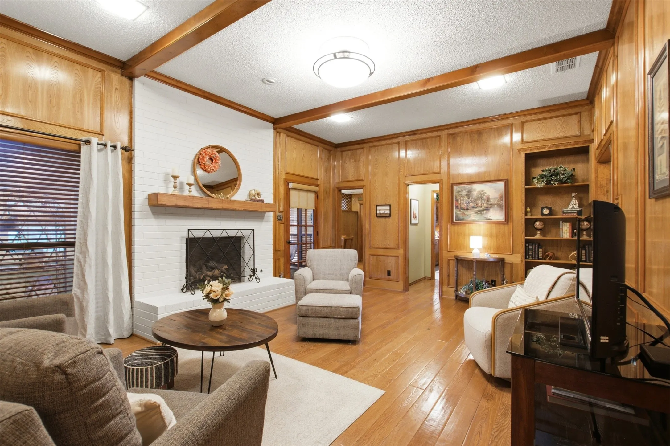 Spacious living area featuring wood-paneled walls, a white brick fireplace with a wood mantel, wood-finish flooring, exposed ceiling beams, and built-in shelving