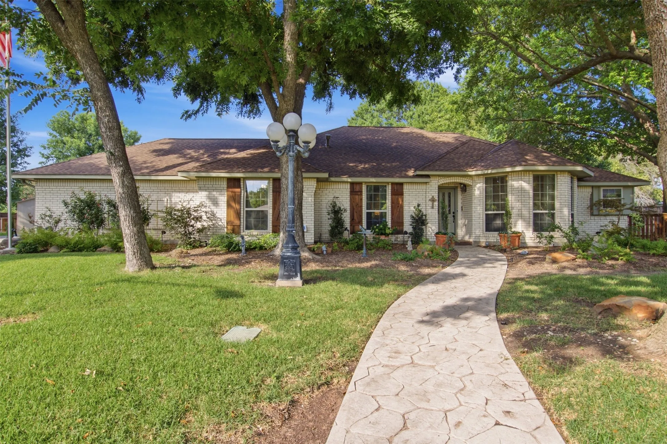 Single-story residence featuring light-colored brick exterior, a stamped concrete walkway, mature trees, a multi-lamp yard light, and decorative window shutters
