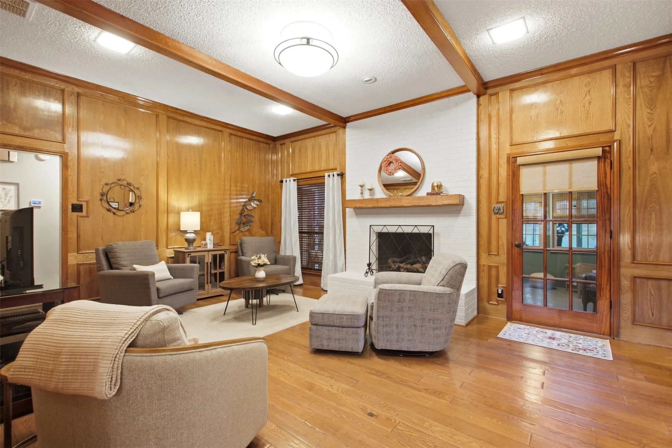 Living area featuring wood-finish flooring, wood-paneled walls, exposed wood ceiling beams, and a white brick fireplace with a wood mantle