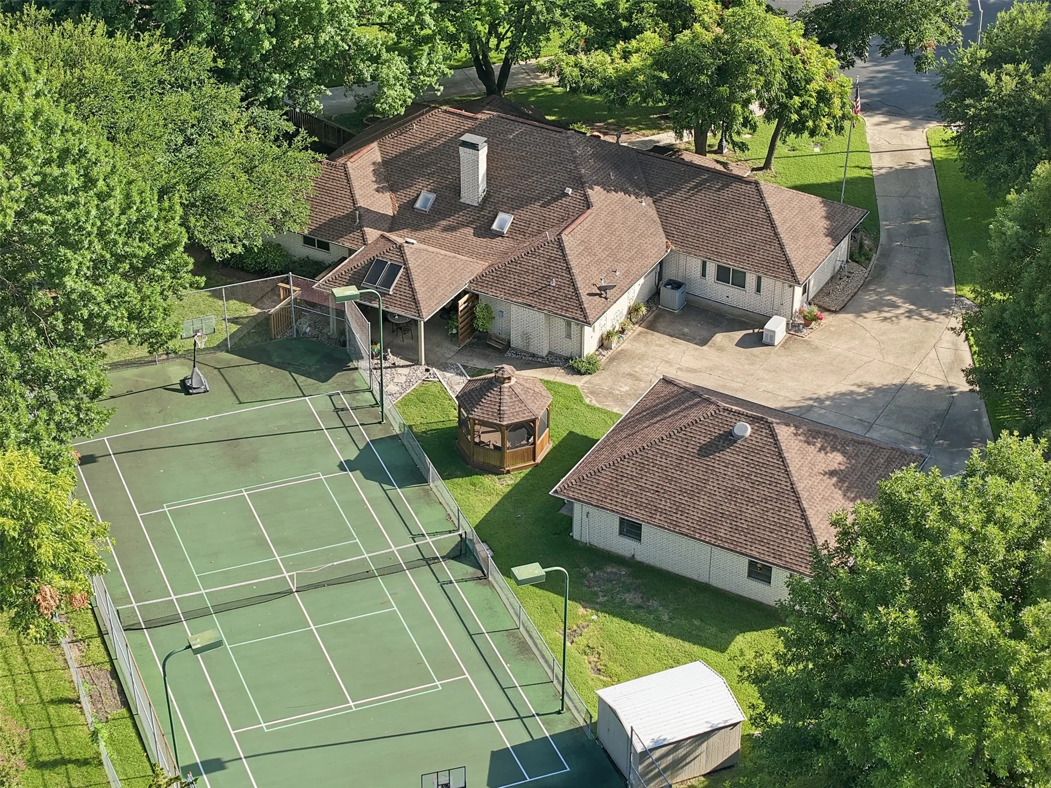 Expansive outdoor recreation area featuring a full-size tennis court with perimeter fencing and lighting, a covered gazebo, and mature tree landscaping