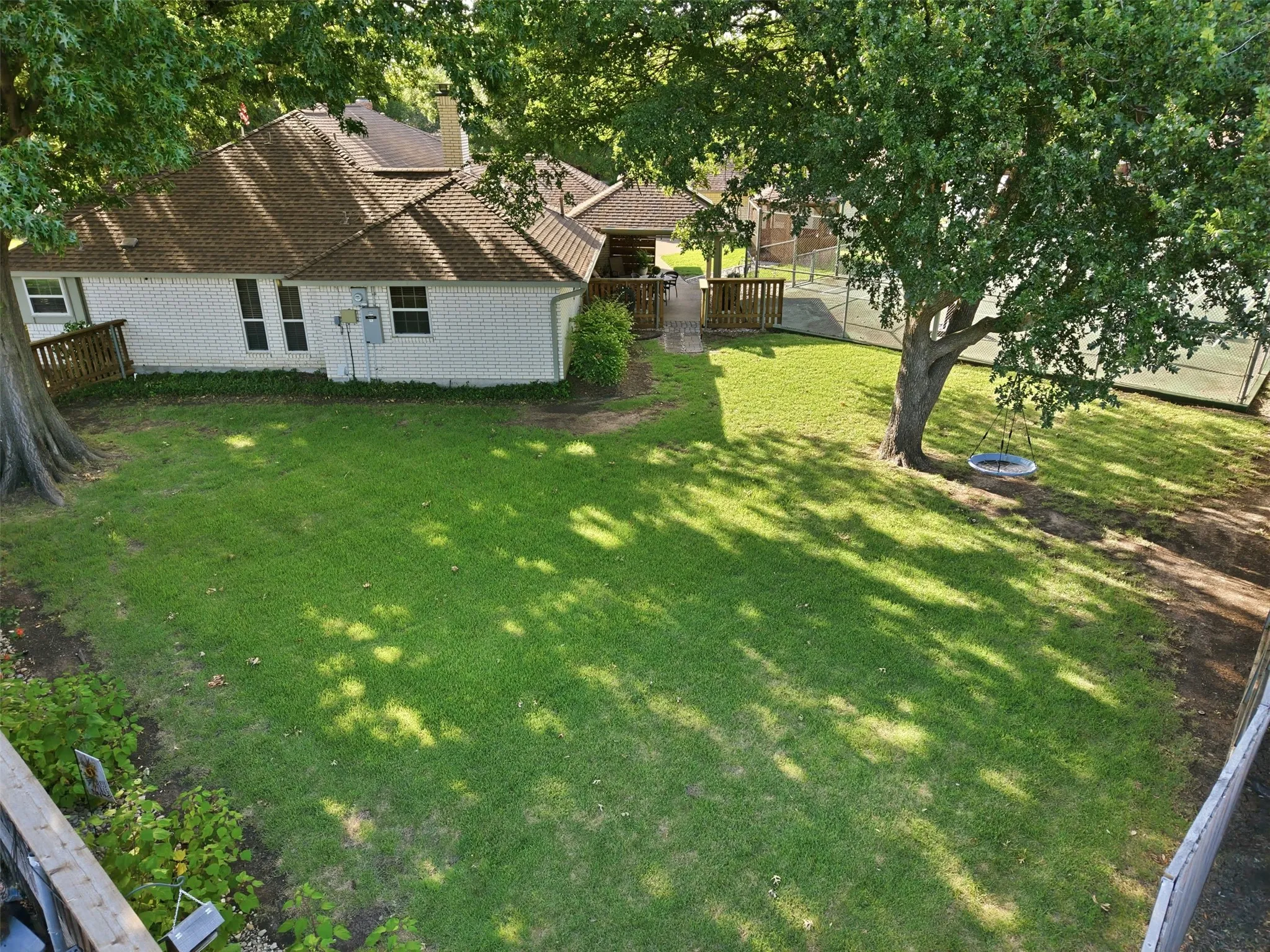 Spacious backyard featuring a manicured lawn, mature trees providing natural shade, a white brick exterior, and an adjacent tennis court
