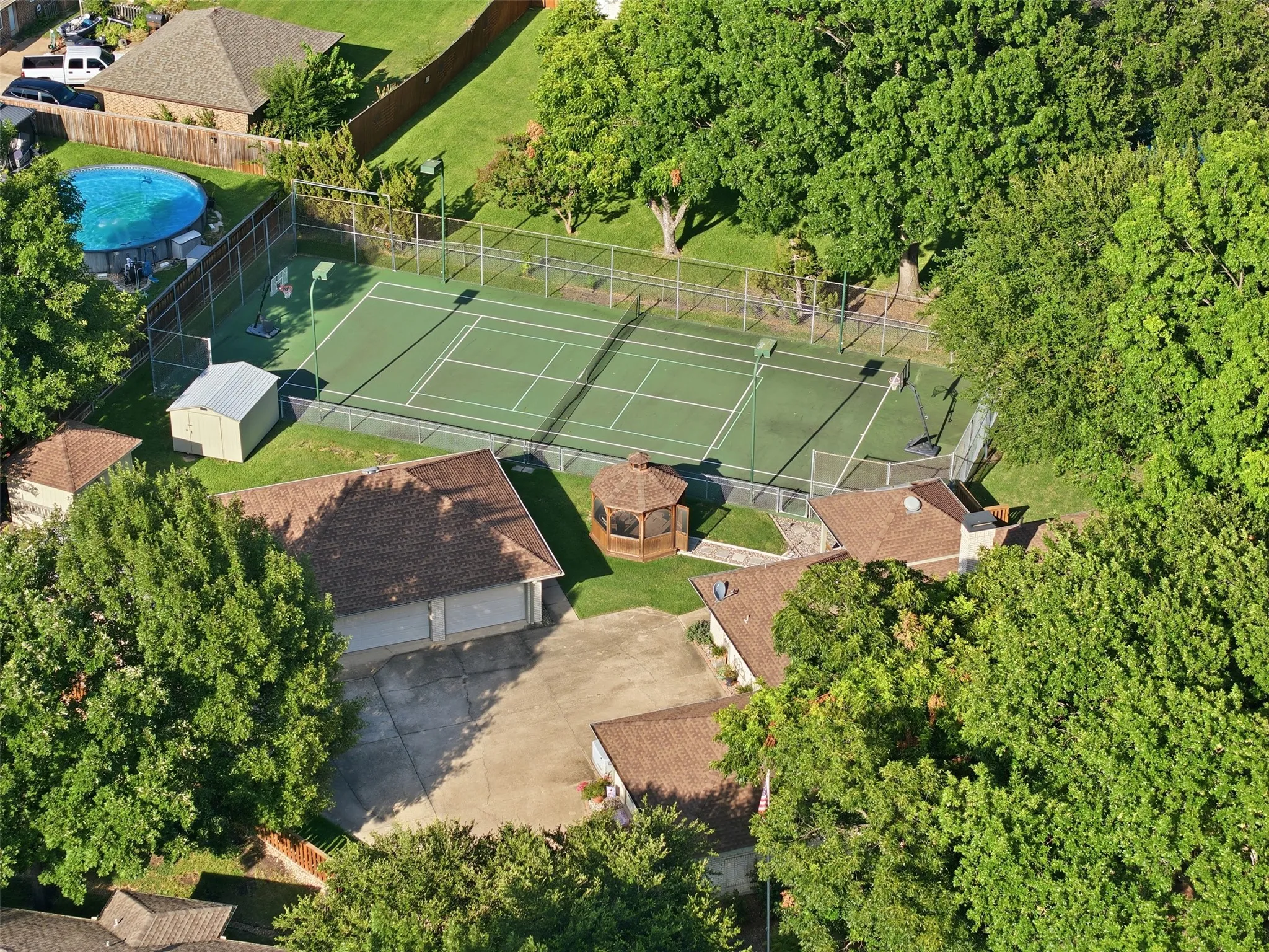 Expansive outdoor recreation area featuring a full-size tennis court, detached gazebo, and mature perimeter trees
