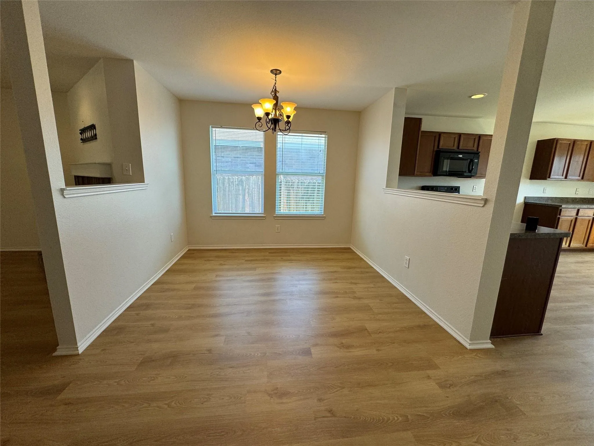 Open-concept dining area featuring wood-finish flooring, a contemporary chandelier, two windows with blinds, and a partial view into the kitchen with dark wood cabinetry and a built-in microwave