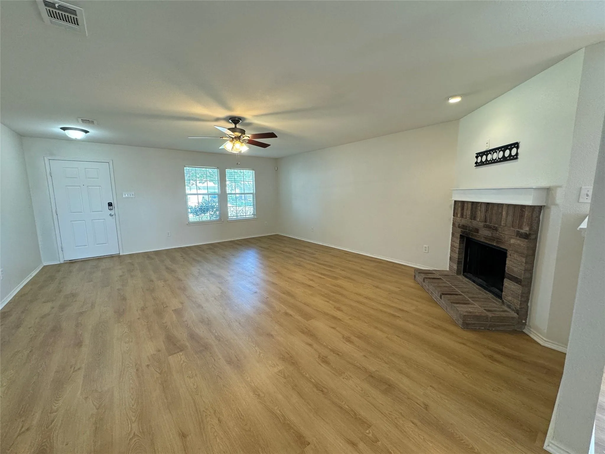 Spacious living area featuring wood-finish flooring, a brick fireplace with a white mantel, and ample natural light from two large windows